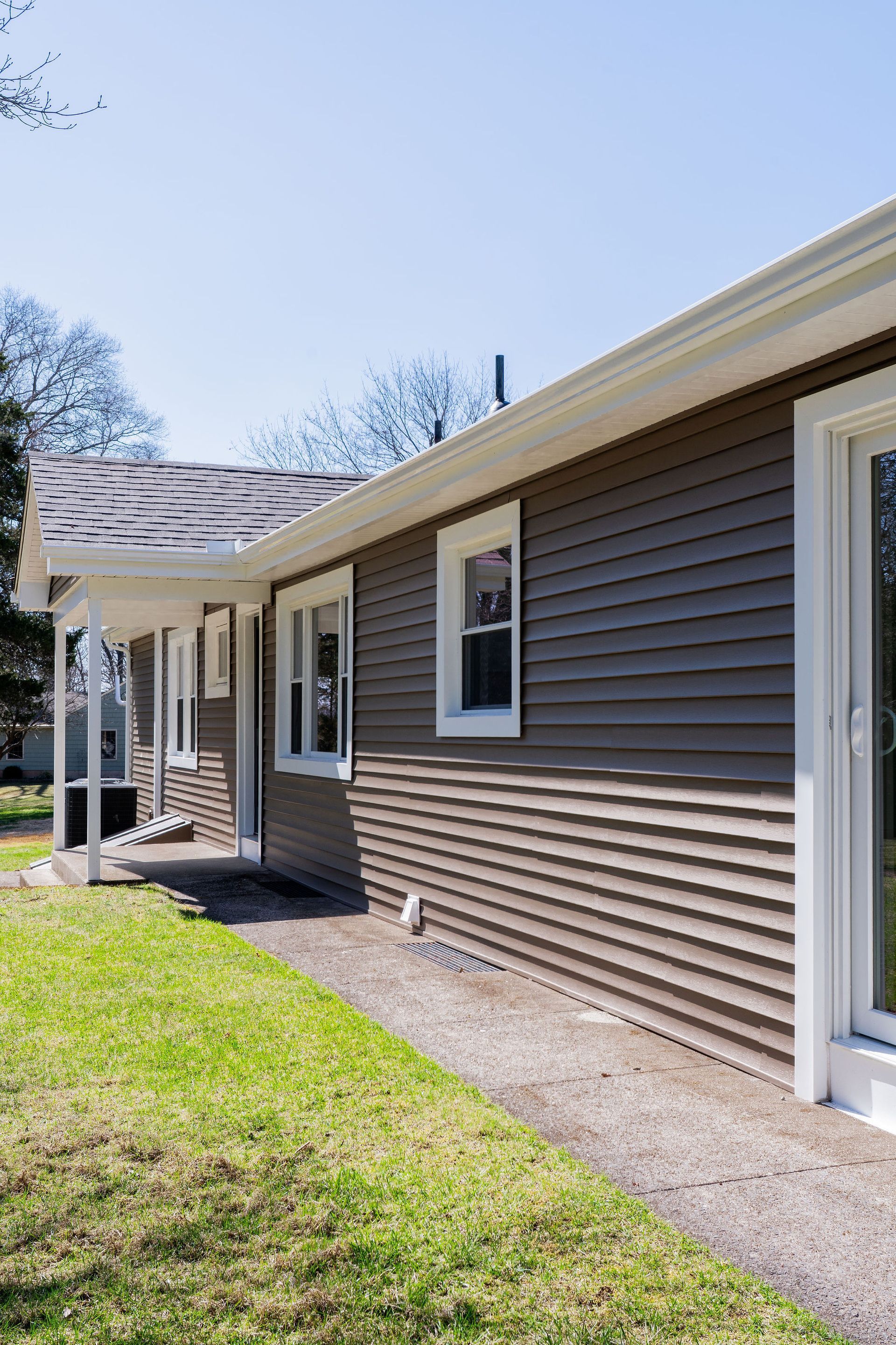 Brown-sided house with white trim, a small porch, and a green lawn under a blue sky.