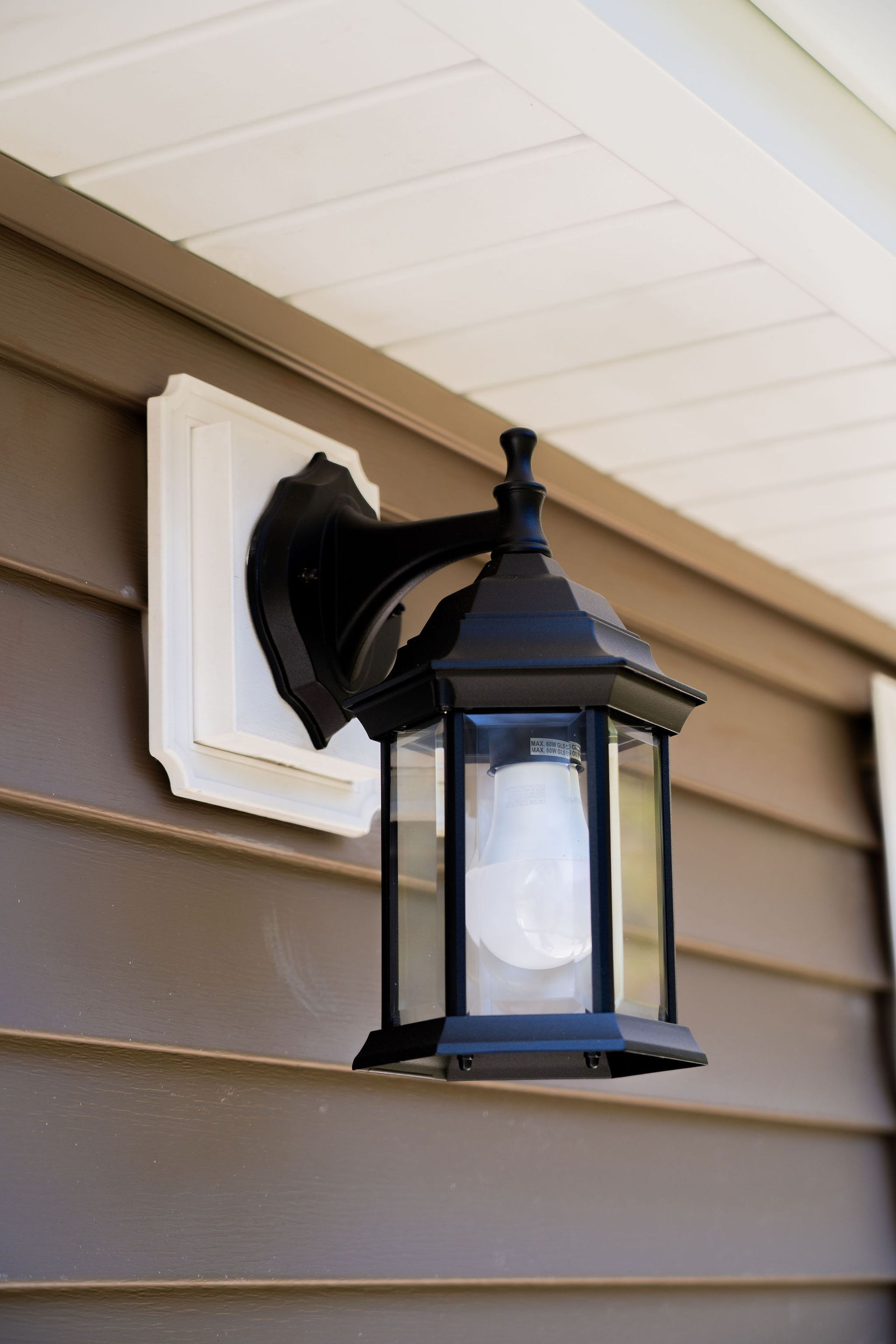 Black outdoor lantern on a brown wall with a white mounting plate, under a white ceiling.