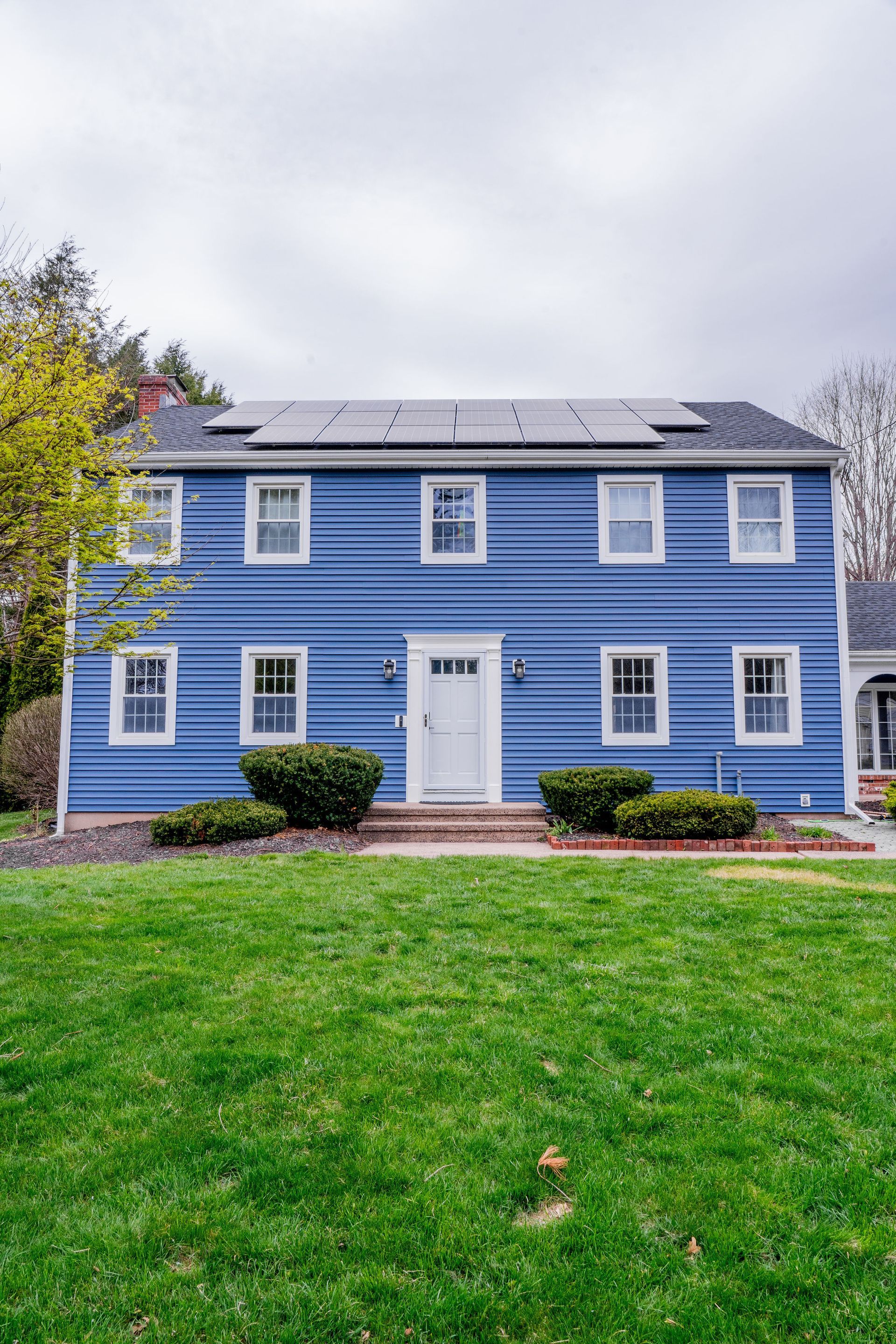 Blue two-story house with white trim, solar panels, and a green lawn under a cloudy sky.