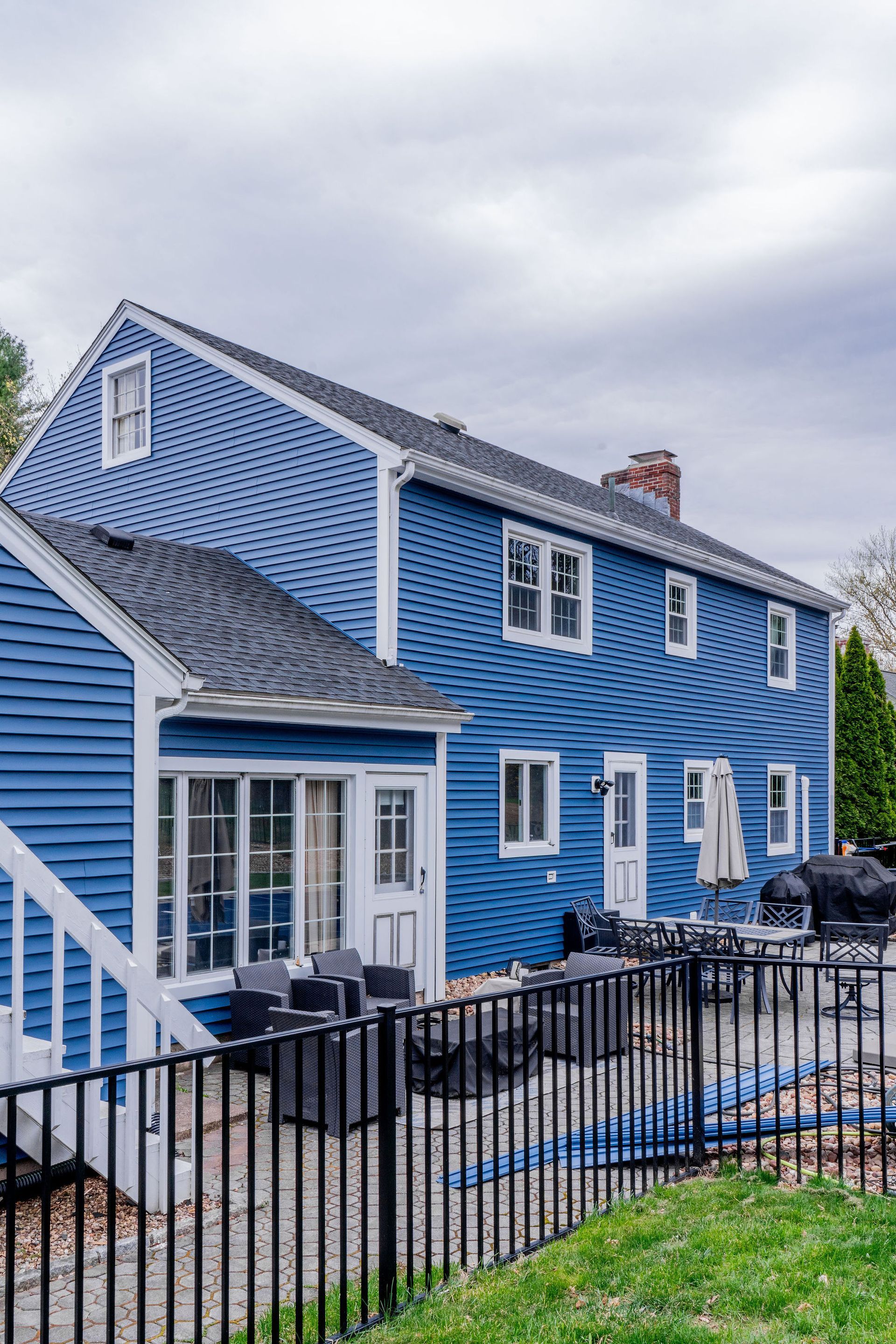 Blue house with white trim, black fence, patio furniture, and a grill. Overcast sky.
