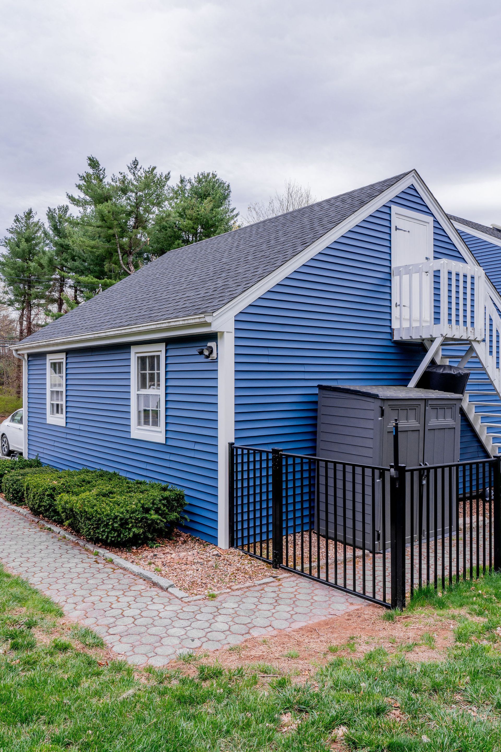 Blue building with white trim, windows, and stairs. Black fence and utility box. Green bushes and grass.