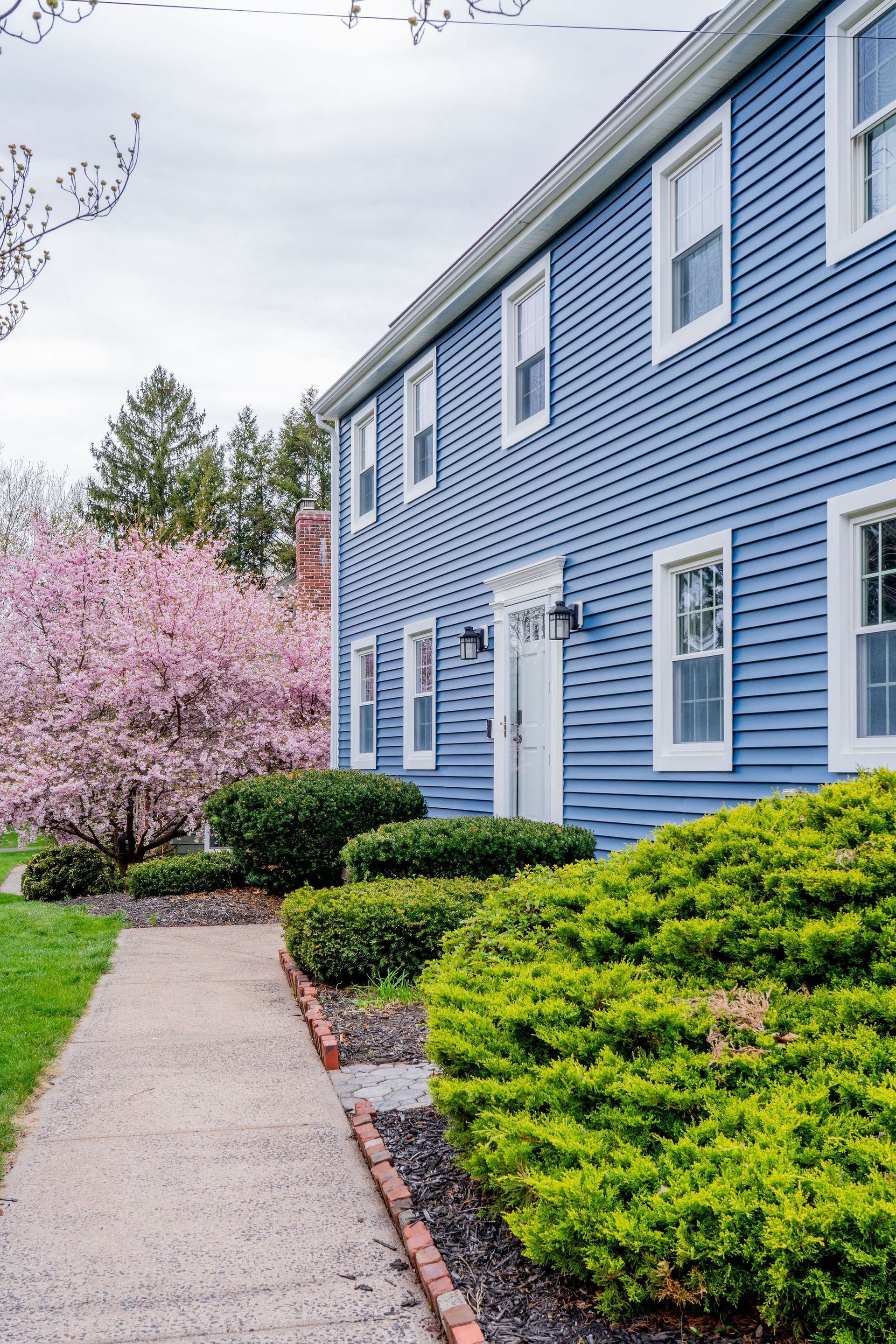 Blue two-story house with white trim and windows; path with shrubbery and pink flowering tree on side.
