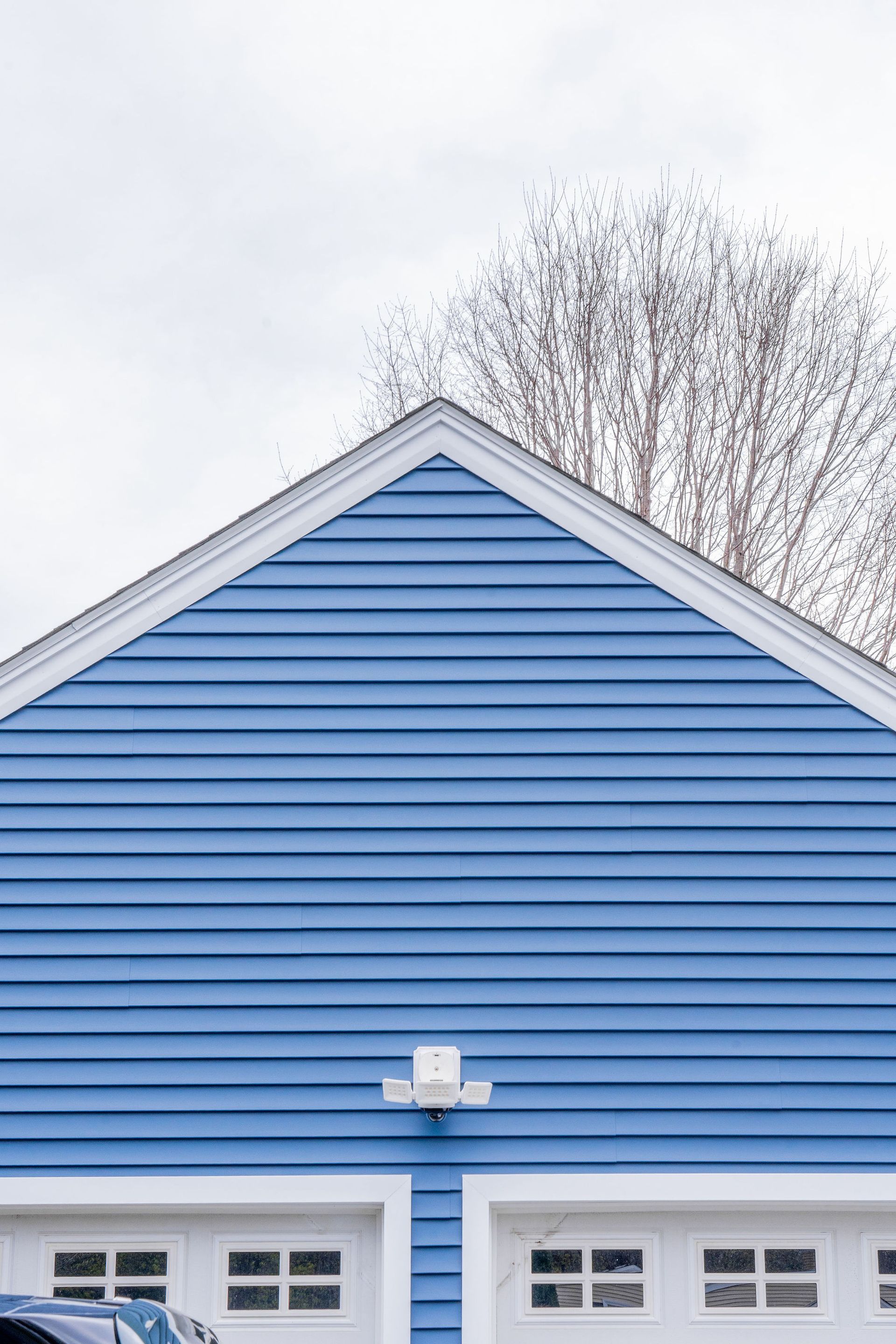 Blue-sided garage with white trim and two garage doors beneath a triangular roof. Tree silhouette against cloudy sky.