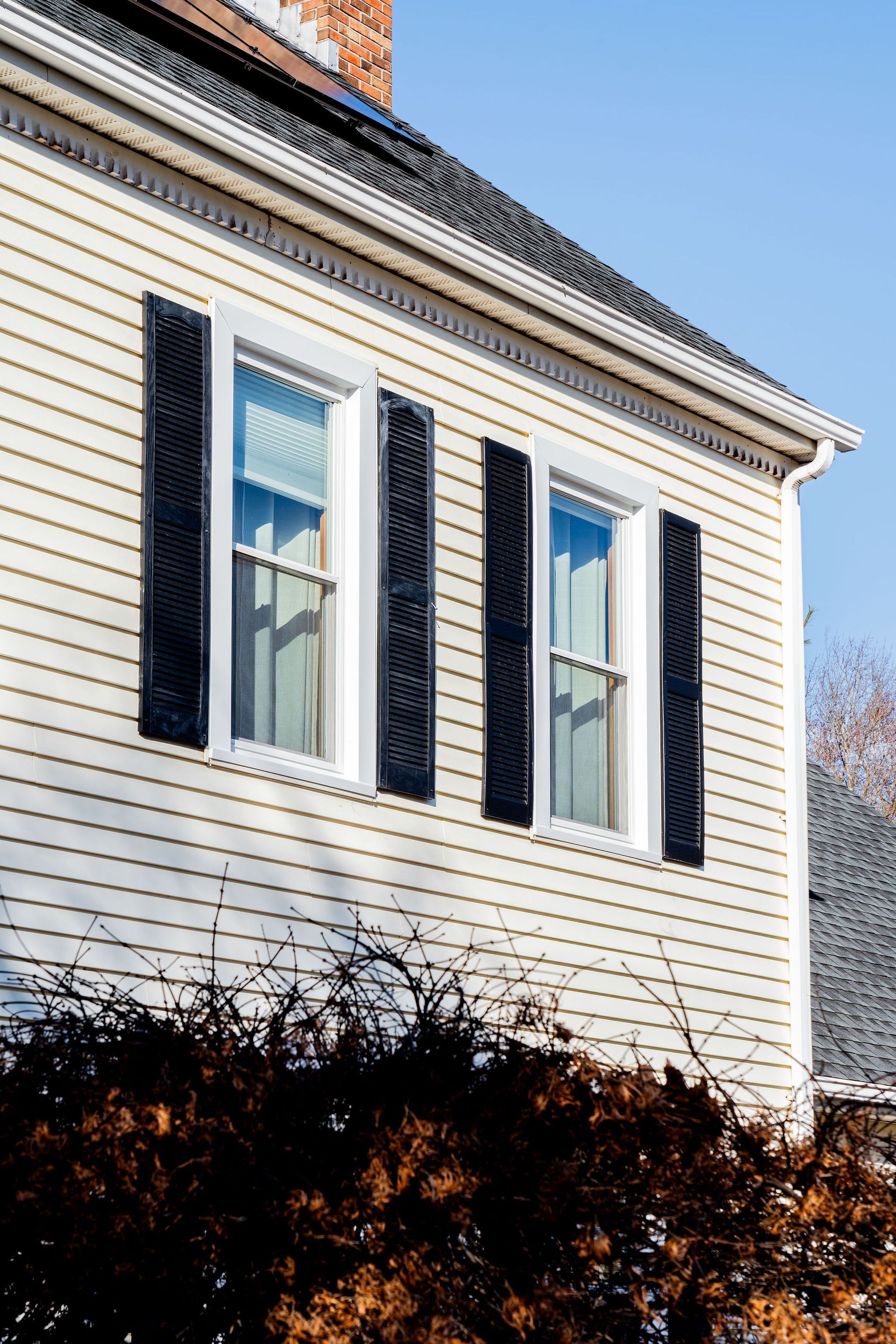 Two windows with black shutters on a light yellow clapboard house, blue sky background.