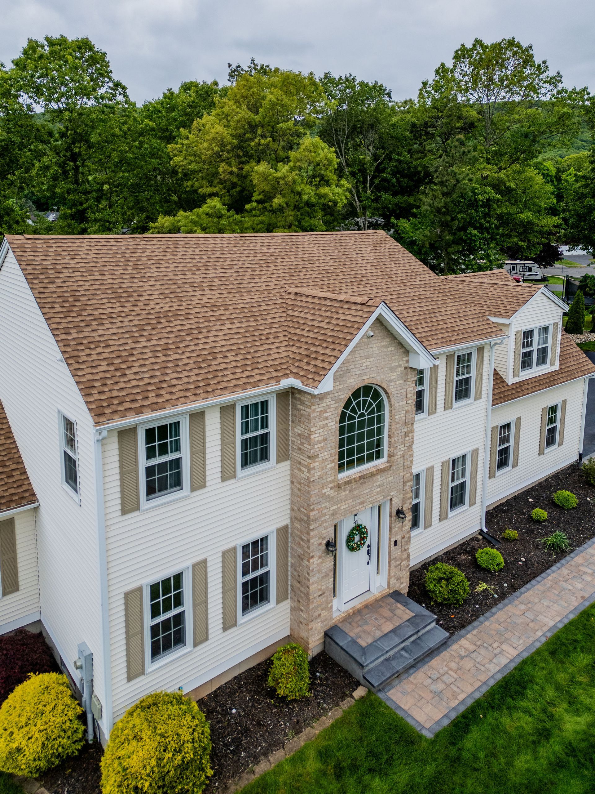 Two-story house with white siding, tan shutters, and brown roof; brick facade, green lawn.