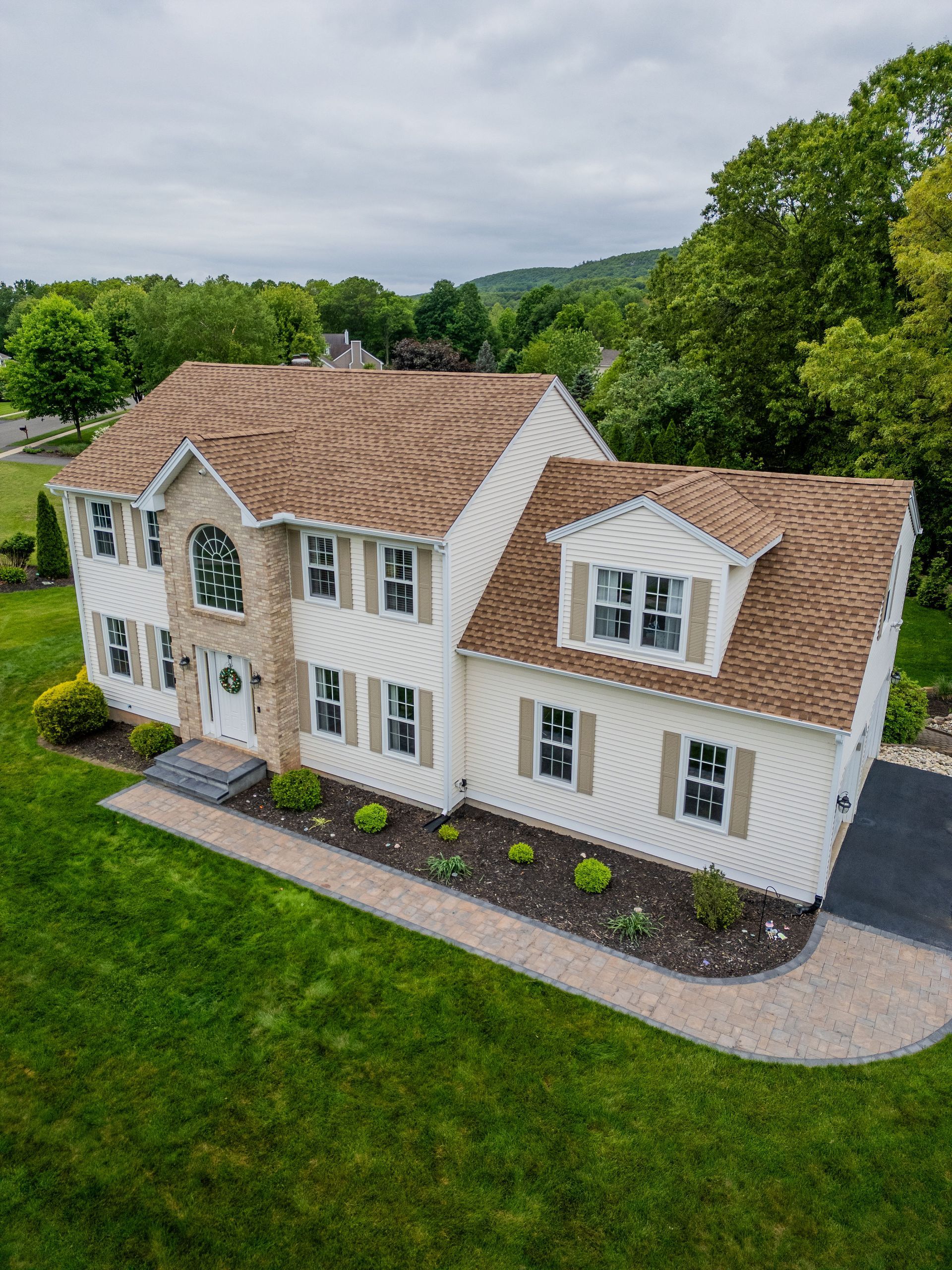 Two-story beige house with brown roof, nestled in greenery. Includes lawn, driveway, and stone pathway.