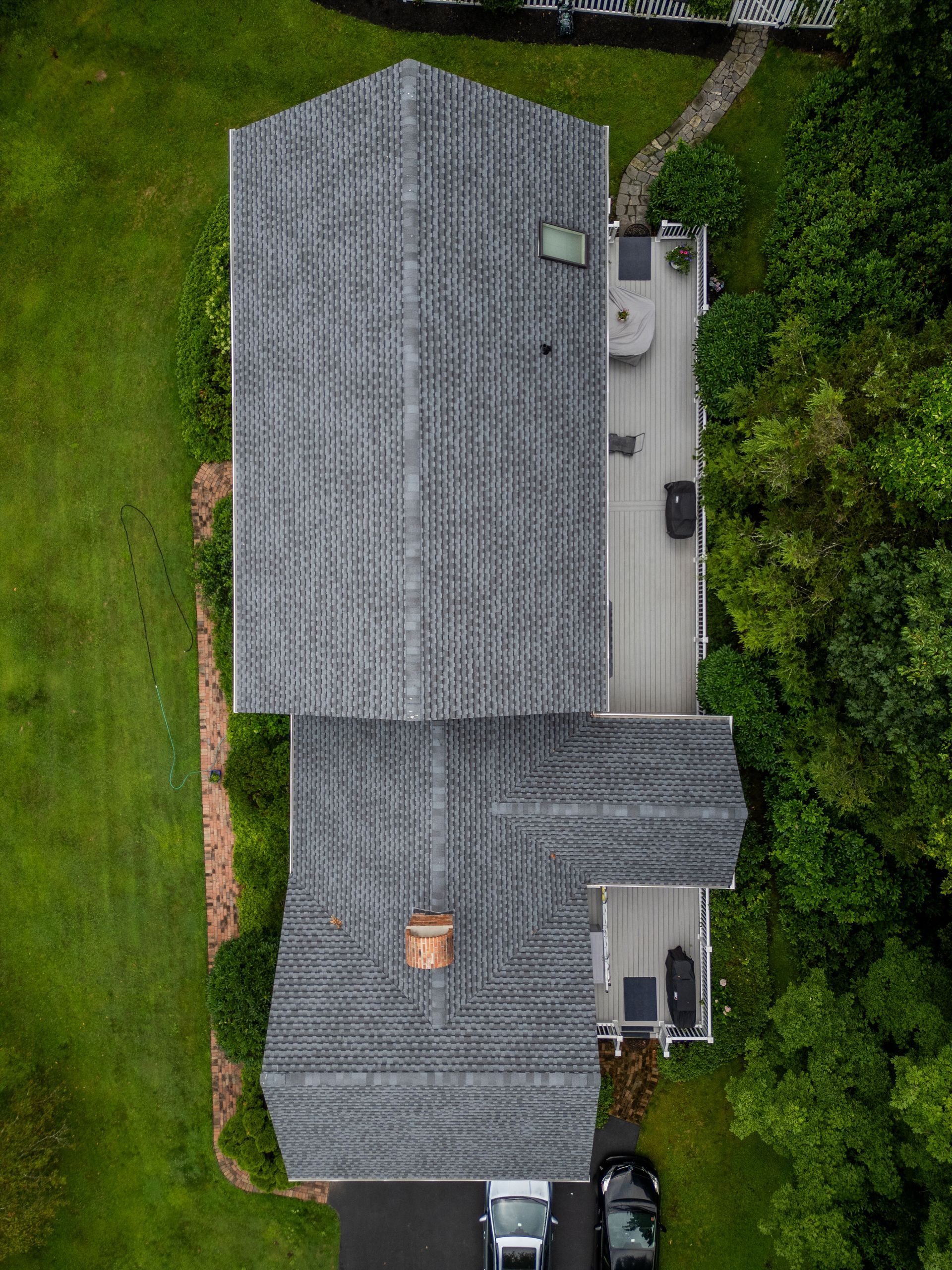 Overhead view of a two-story house with gray roof, deck, and surrounding green lawn and trees.