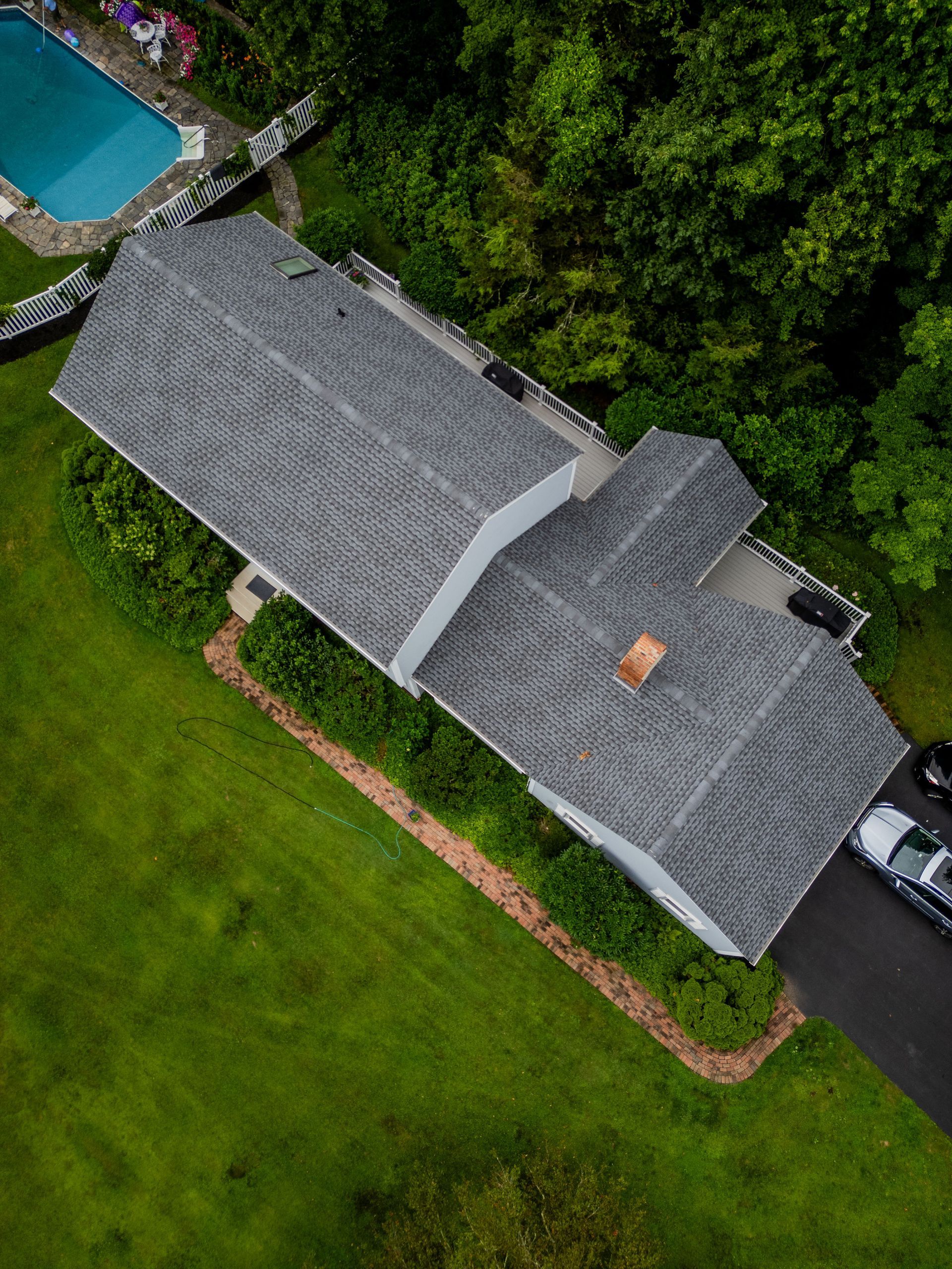 Aerial view of a house with a gray roof, surrounded by green grass, trees, and a pool.