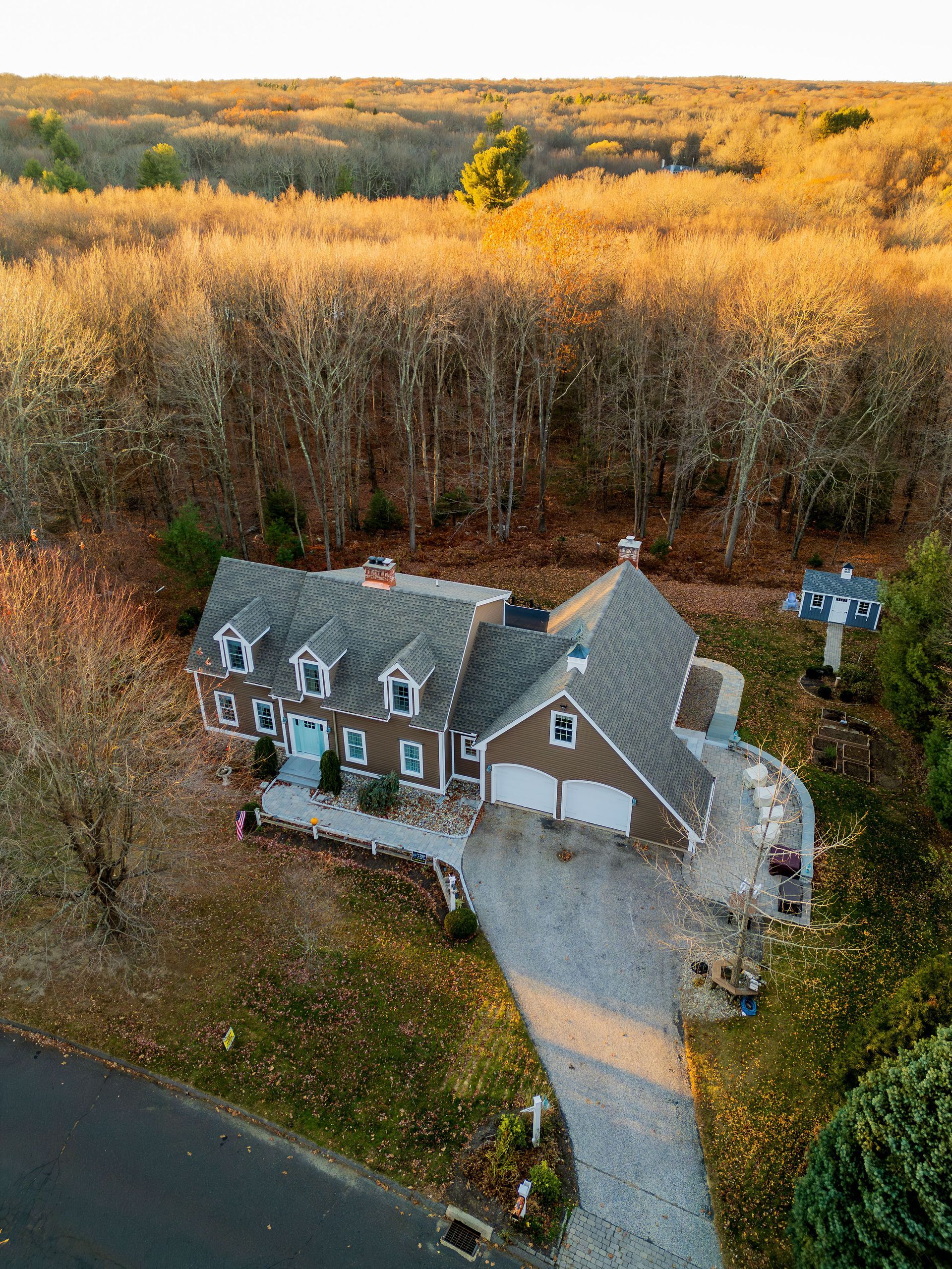Brown house with a gray roof, driveway, and surrounding trees in fall foliage.