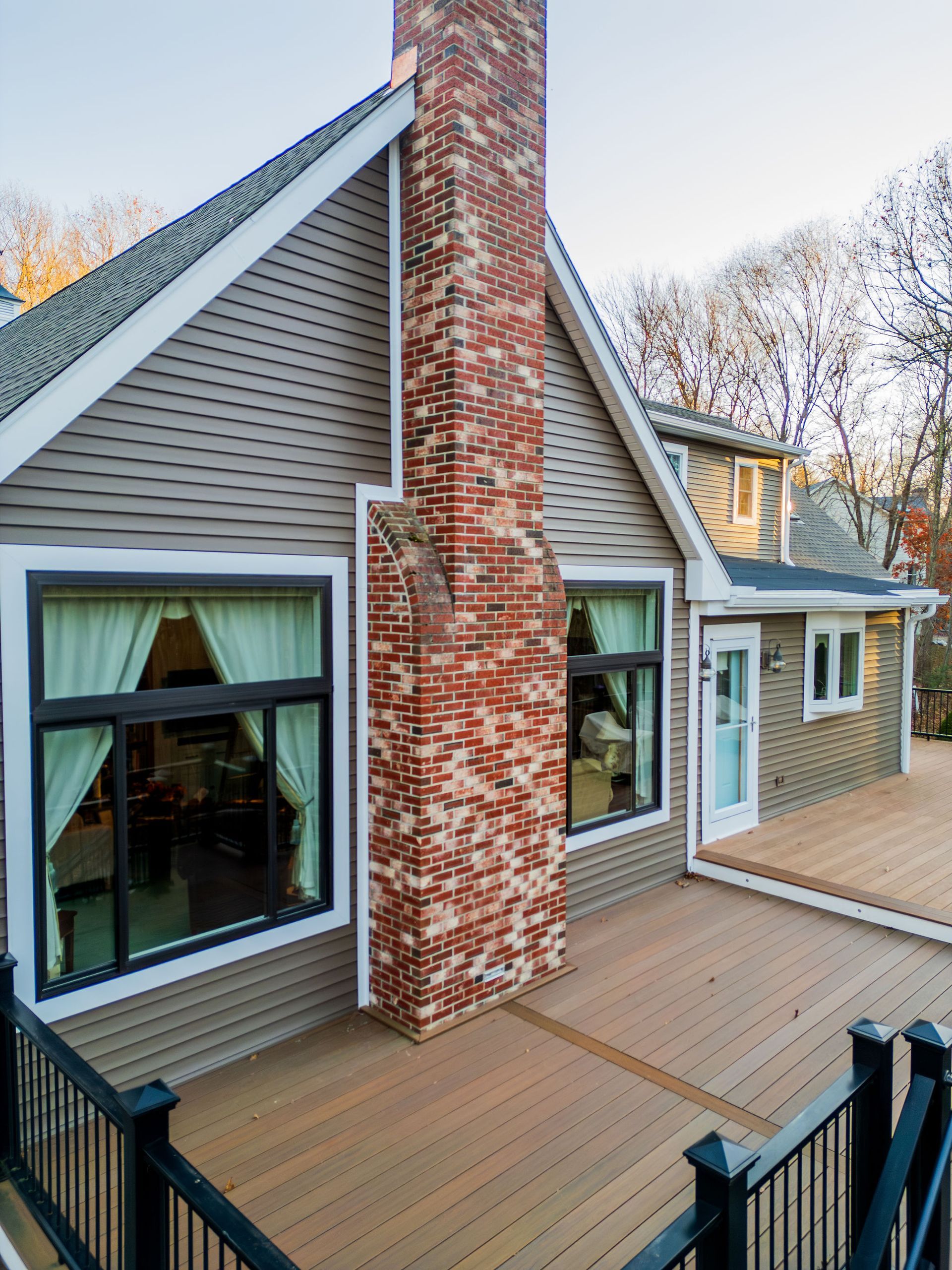 House exterior with brick chimney, brown siding, large windows, and a wooden deck with black railing.