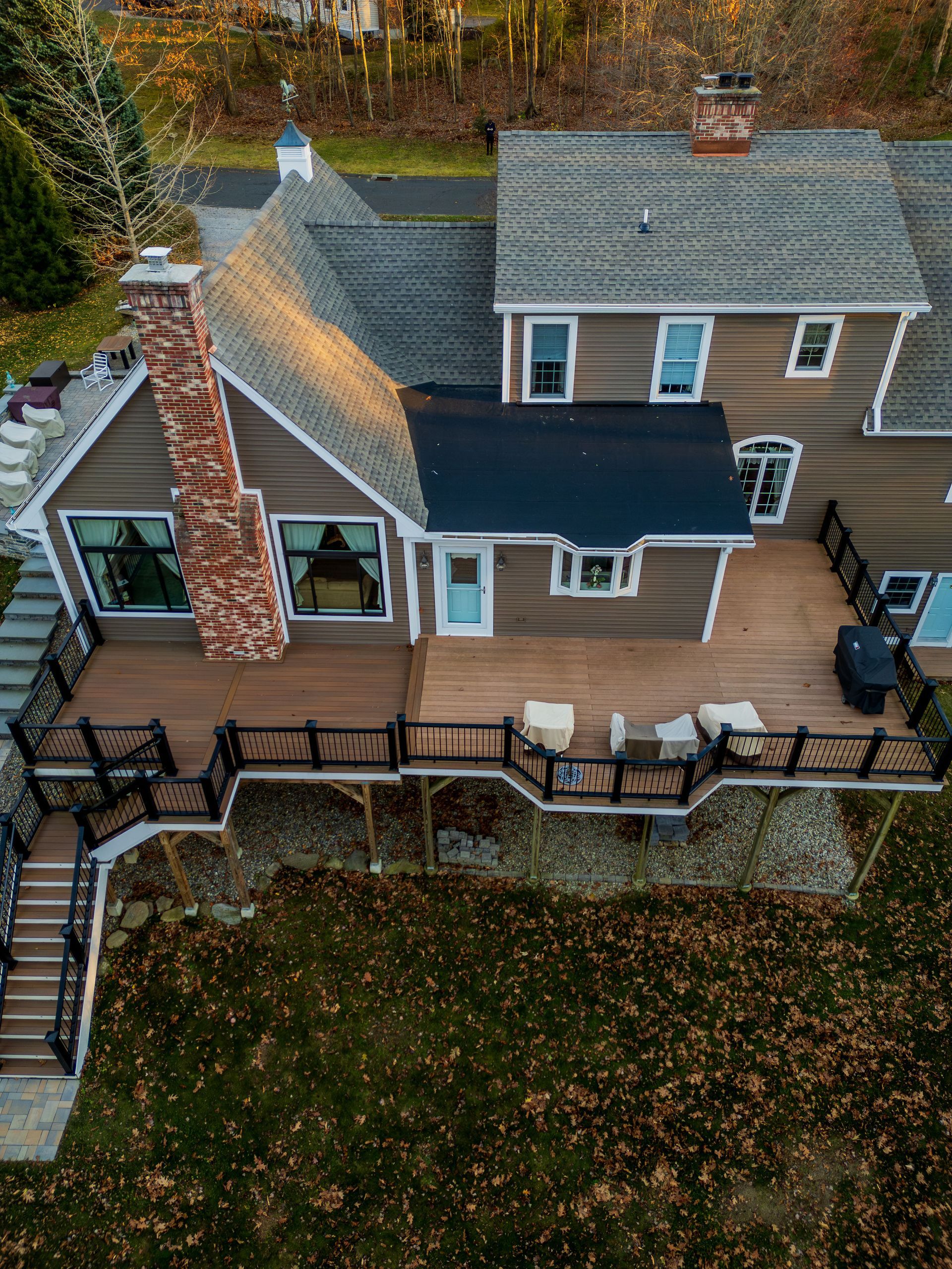 Aerial view of a brown house with a wooden deck, stone staircase, and brick chimney surrounded by foliage.