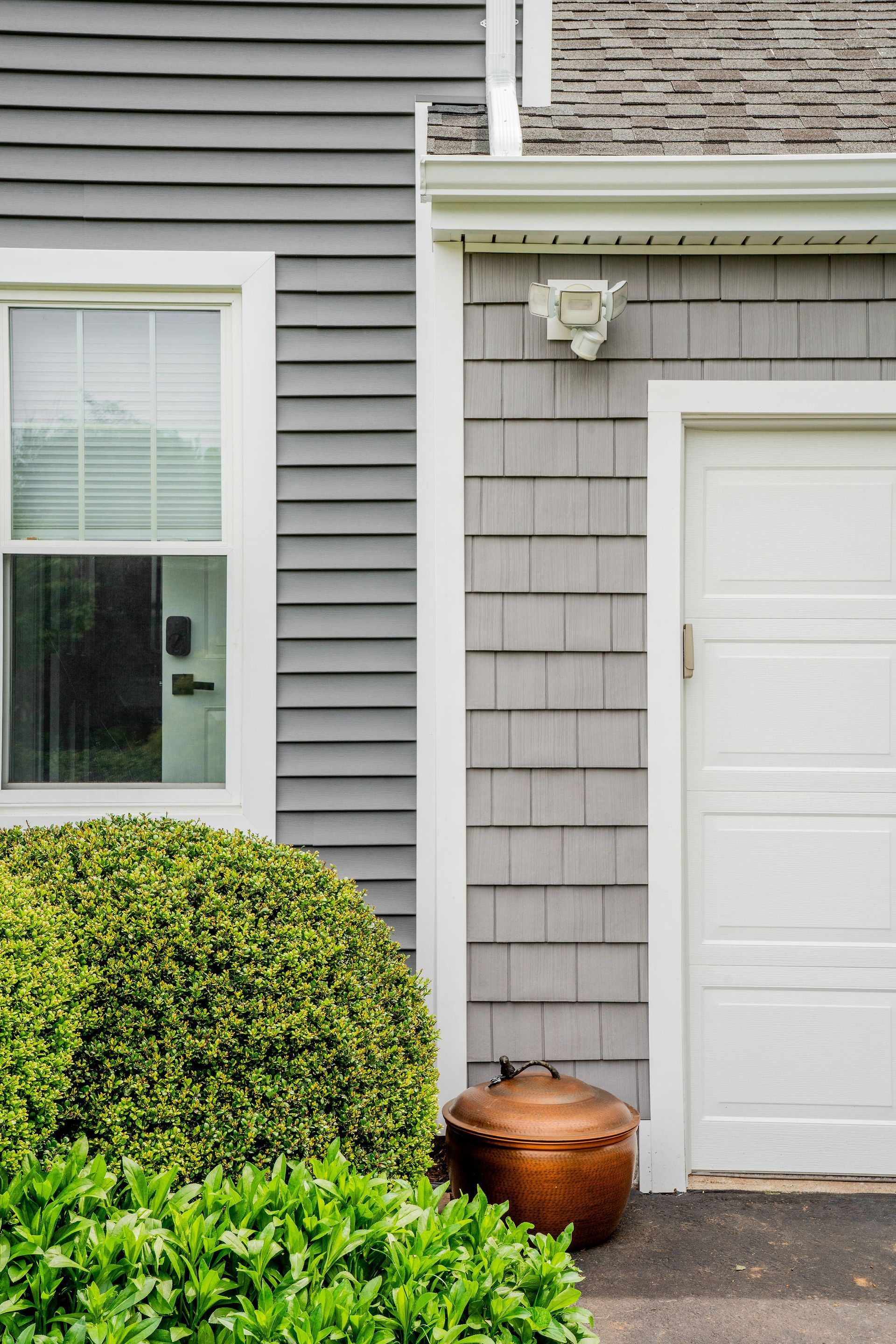 Gray siding and shingle exterior with white-framed window and garage door; a bronze container sits near green shrubbery.