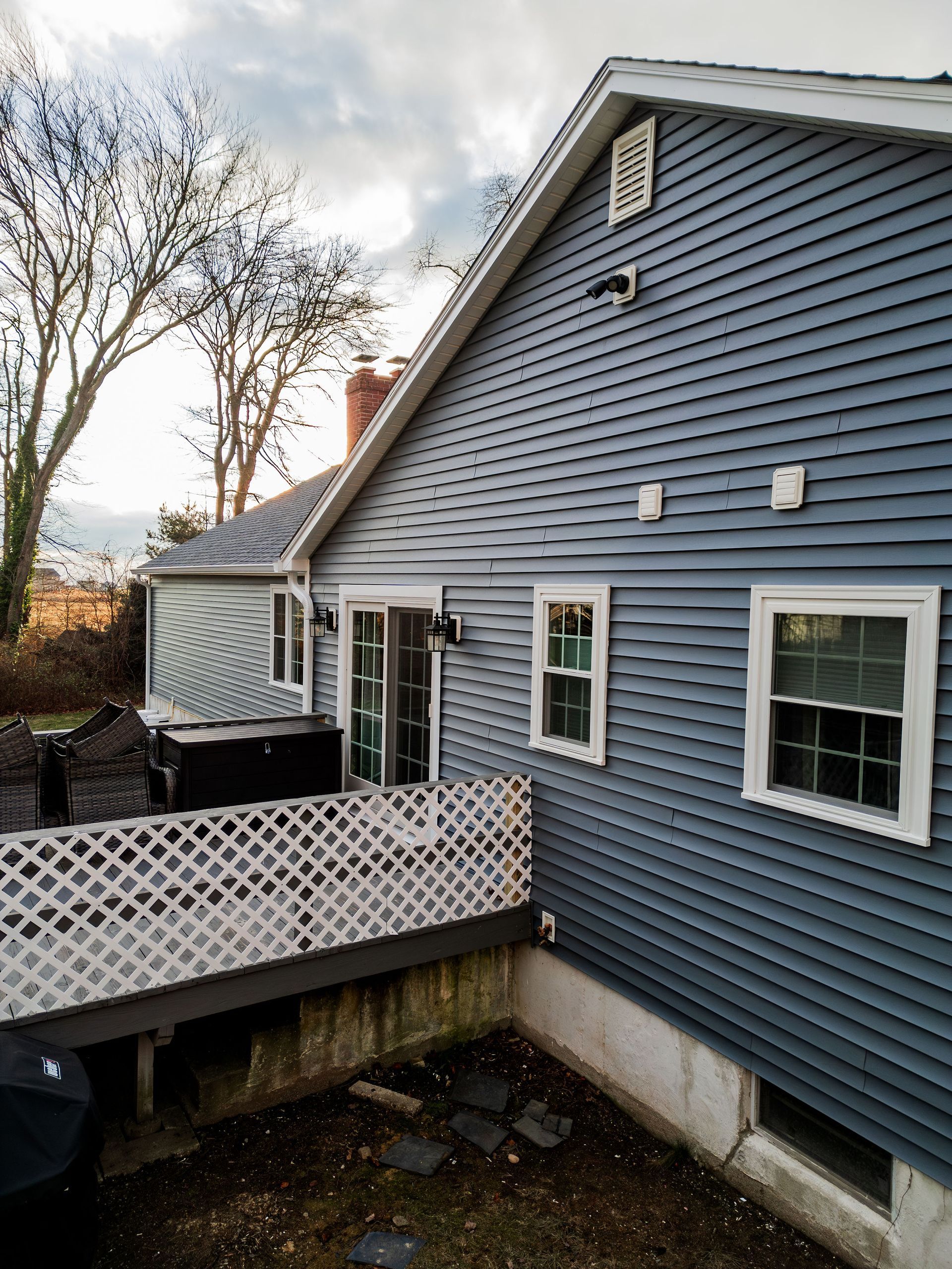 Blue-sided house with white trim and lattice railing in backyard setting. Overcast sky.
