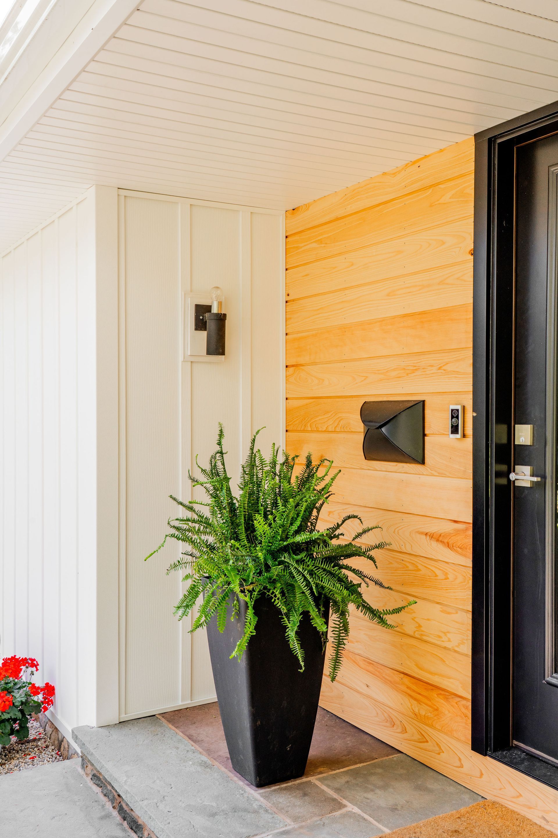 Entryway: Black door, light wood wall, black planter with fern, wall sconce.