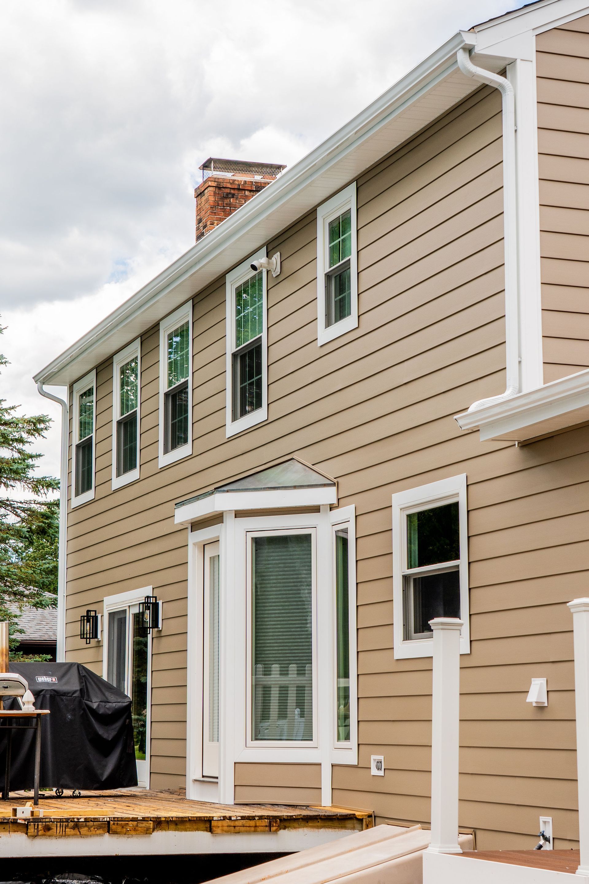Tan house with white trim, windows, deck, and grill.  Cloudy sky.