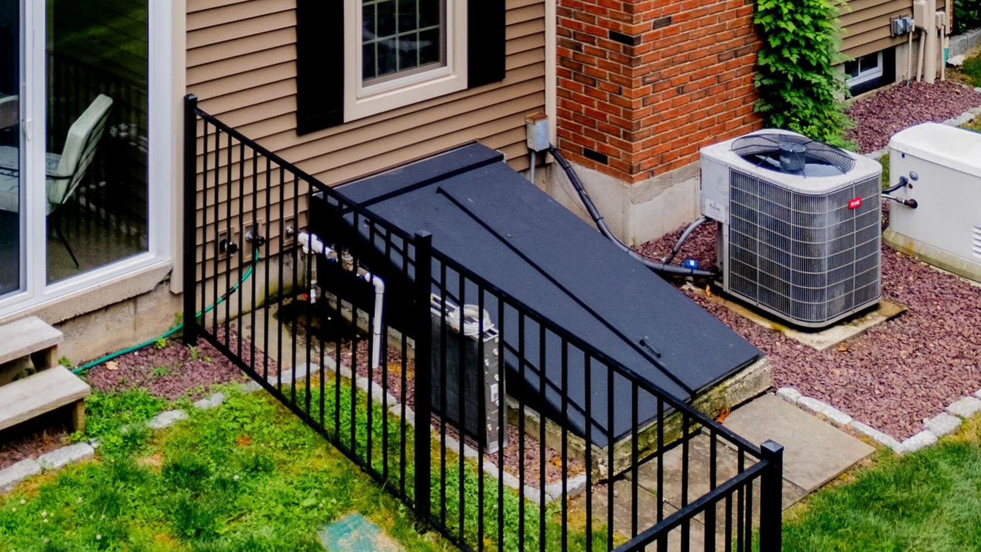 Black fence encloses a black hatch next to an air conditioning unit against a brown building.