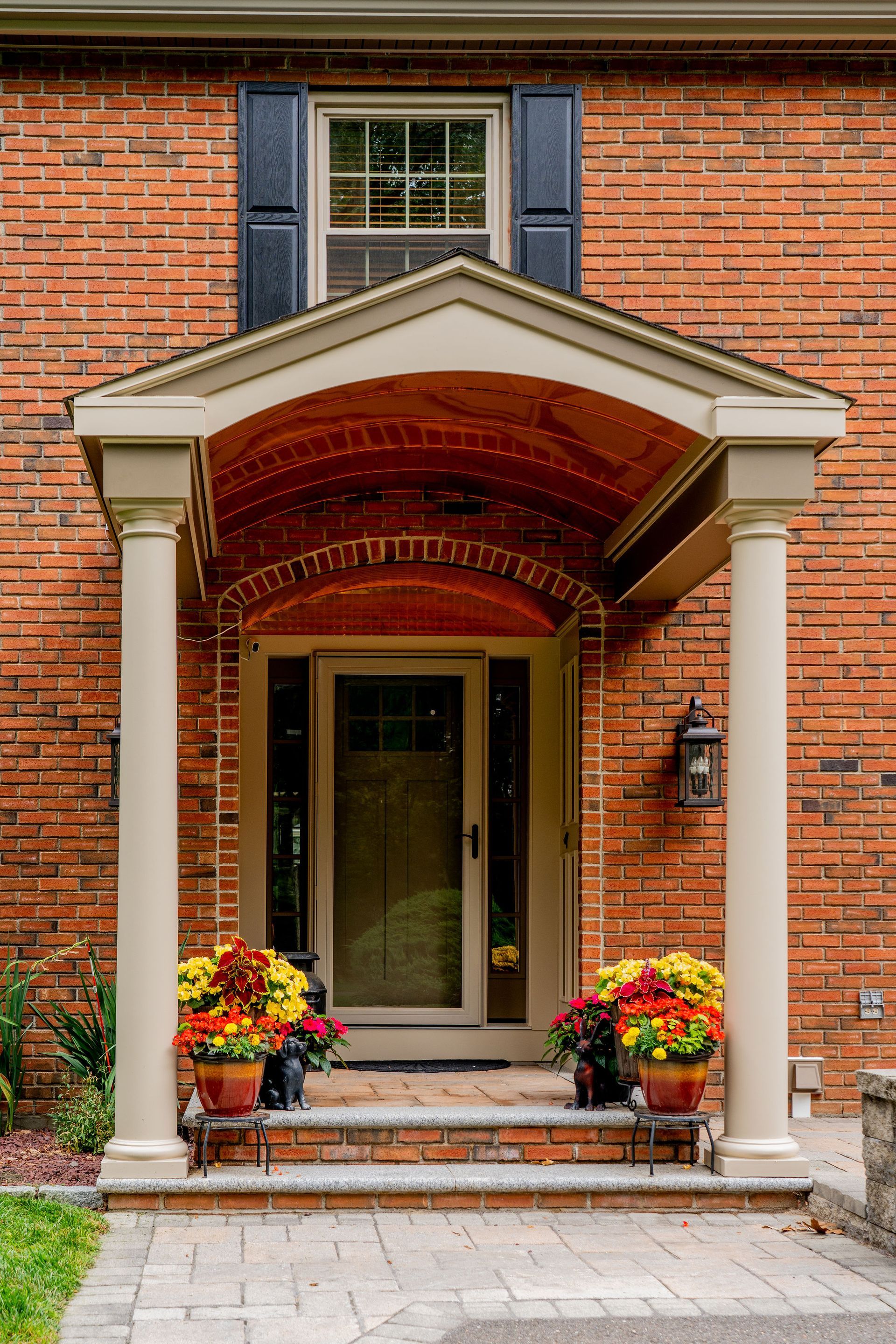 Brick house entry with covered porch, white columns, glass door, and potted flowers.