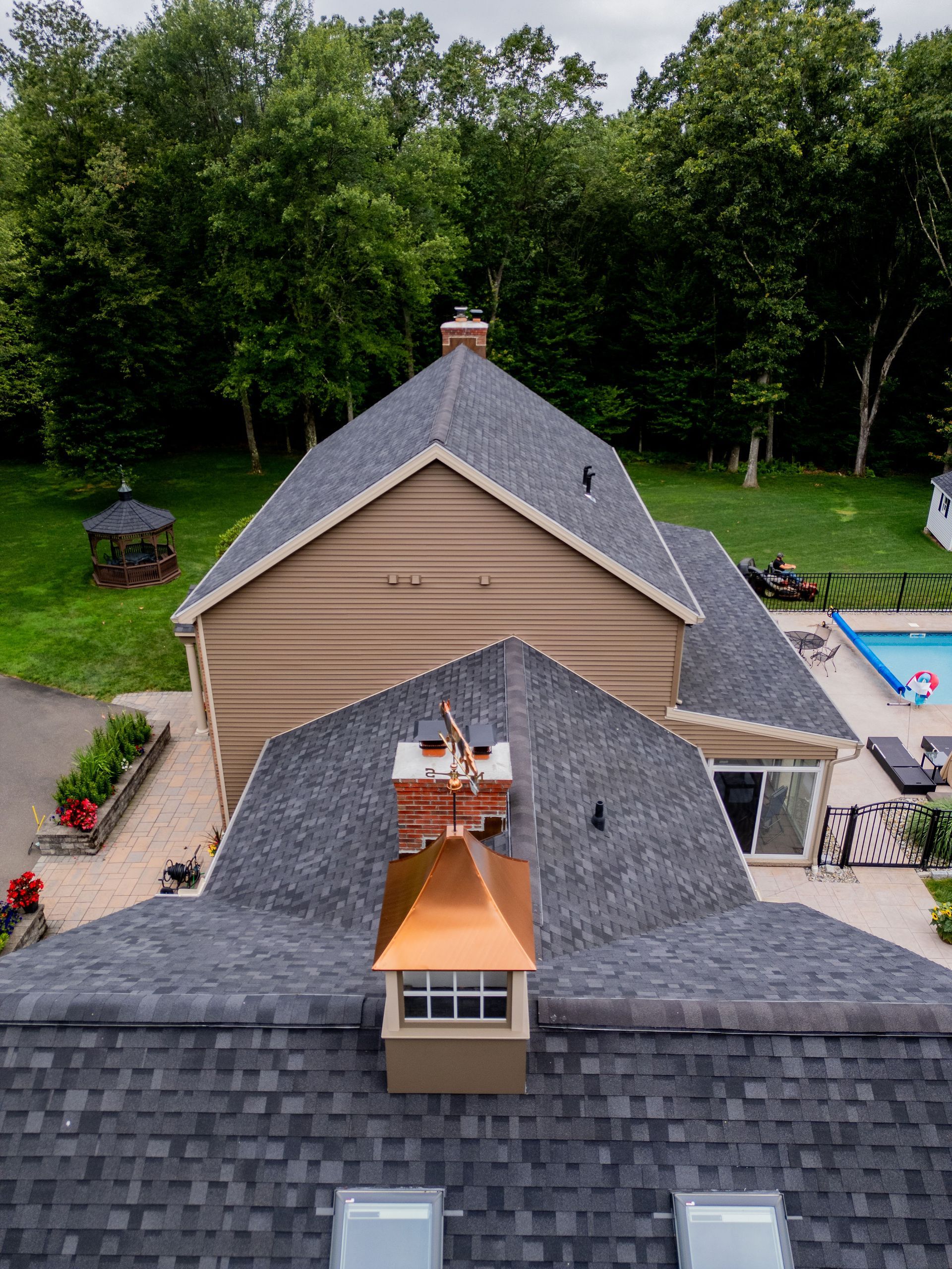 Overhead view of a house with gray shingle roof, chimney, and copper cap. Green lawn and trees in the background.
