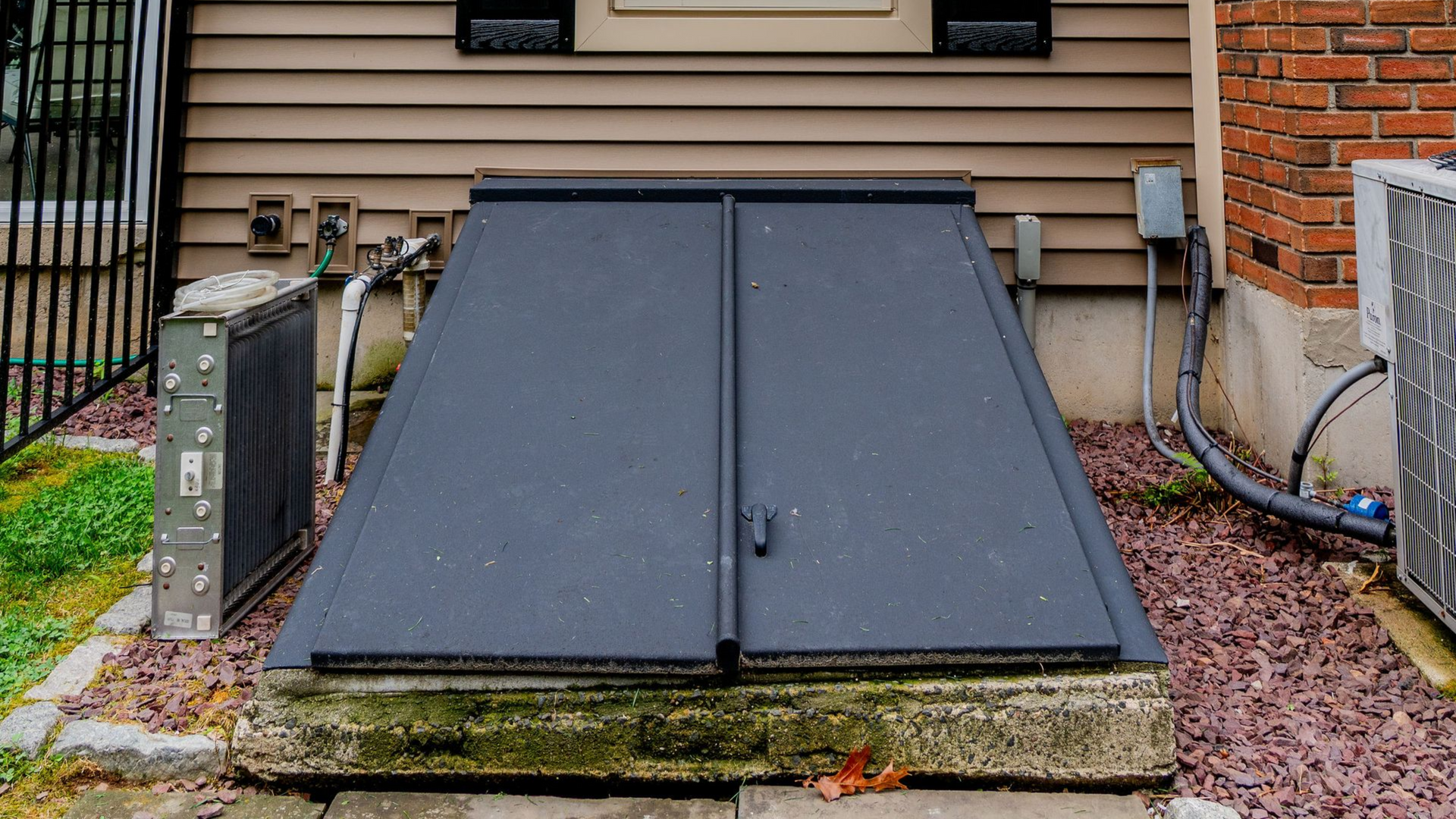 Black metal basement doors on concrete base, beside house with siding and brick, gravel ground.