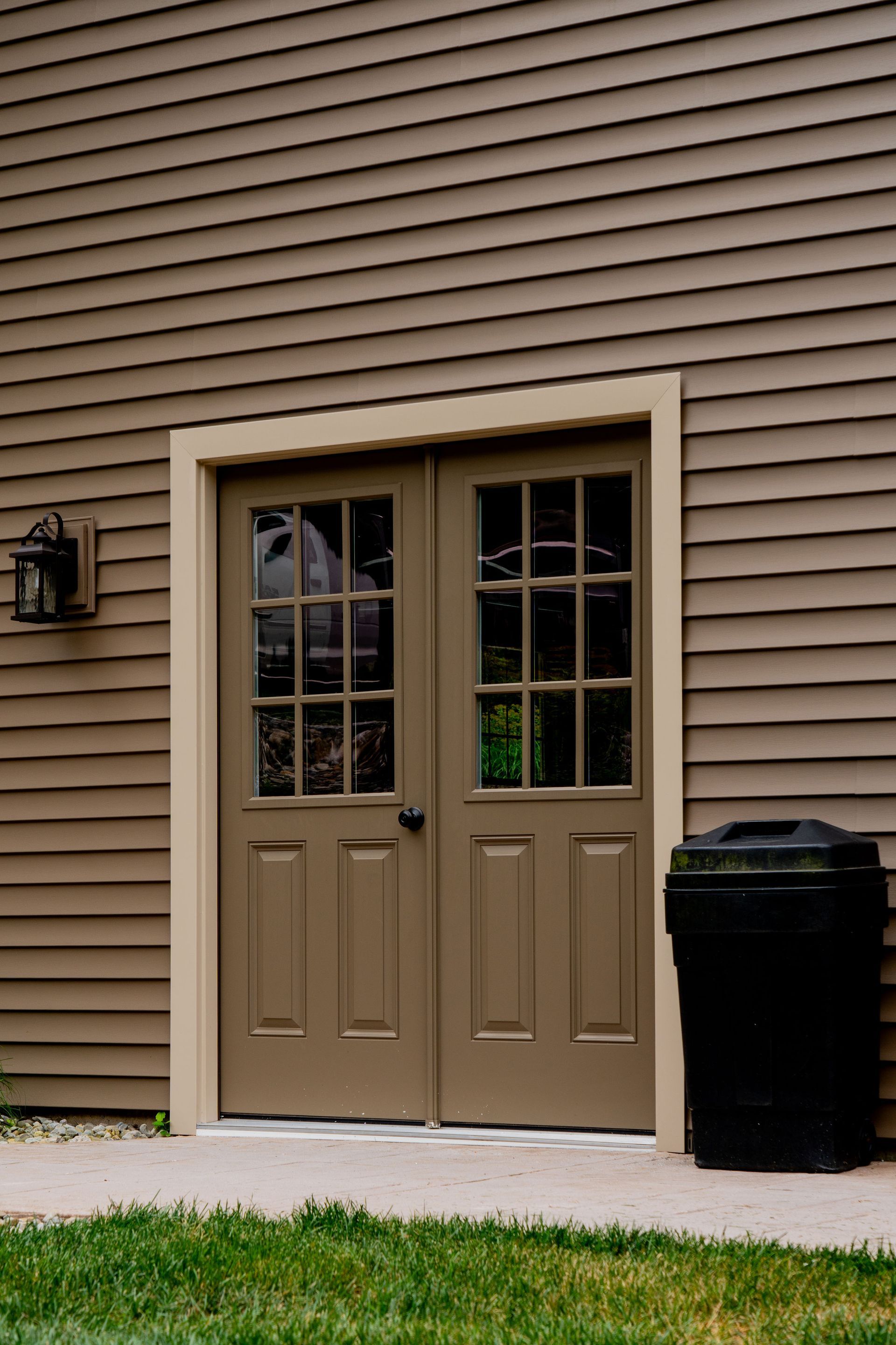 Brown double doors with glass panels and a black trash can next to it, set against a brown clapboard wall.