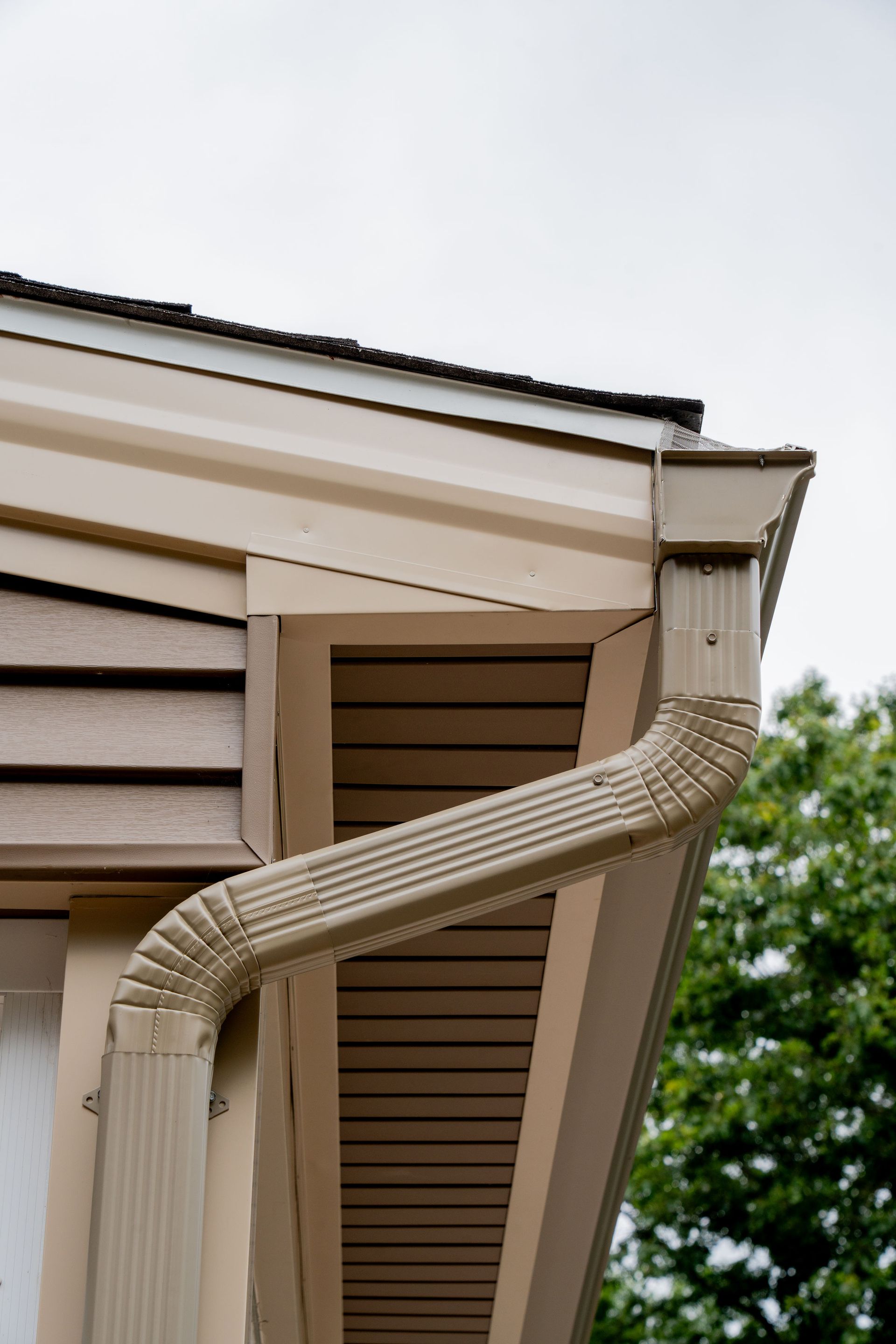 Beige house corner with gutter and downspout against a cloudy sky.