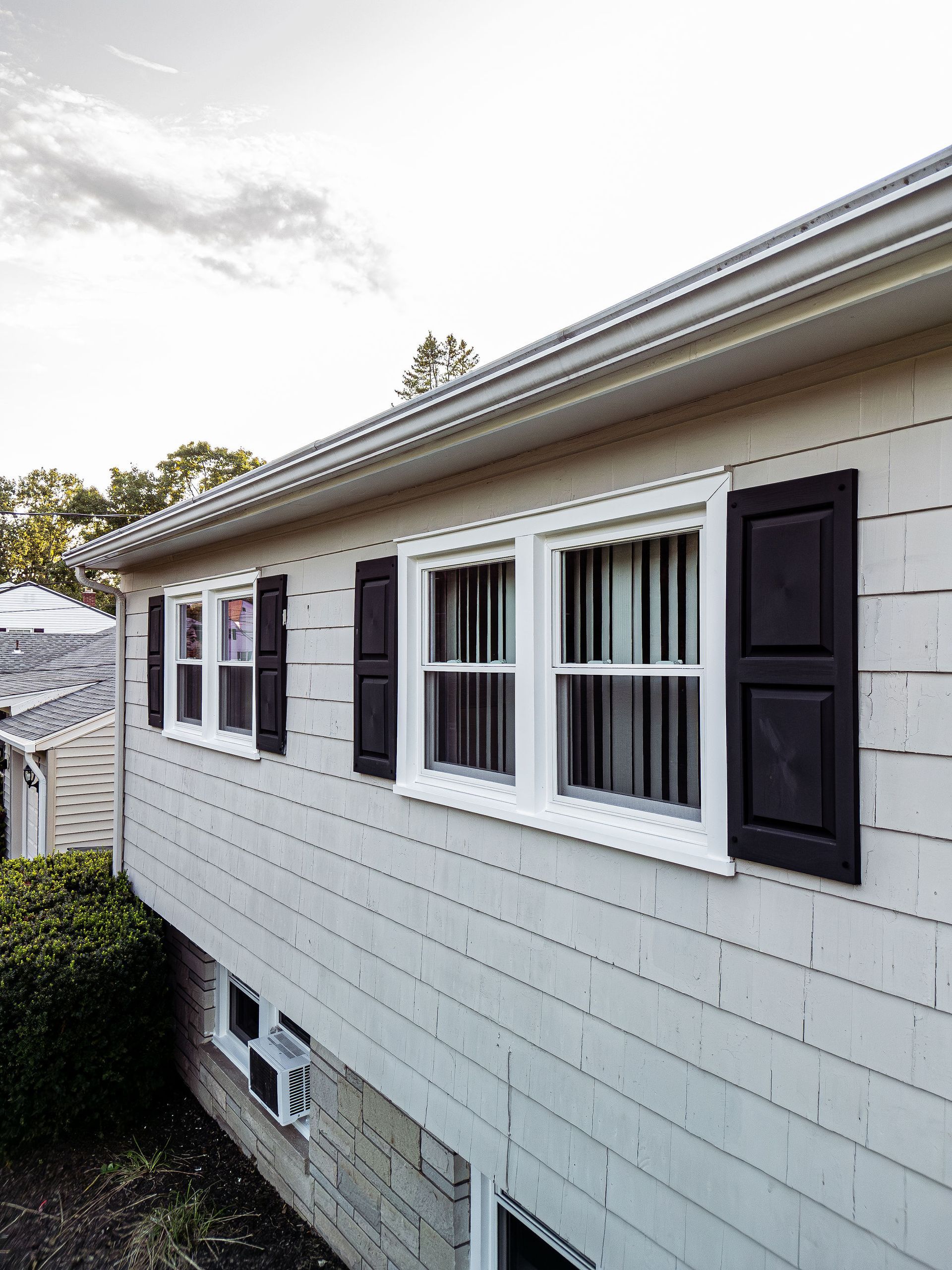 Beige house with white-framed windows and black shutters, gray roof, overcast sky.