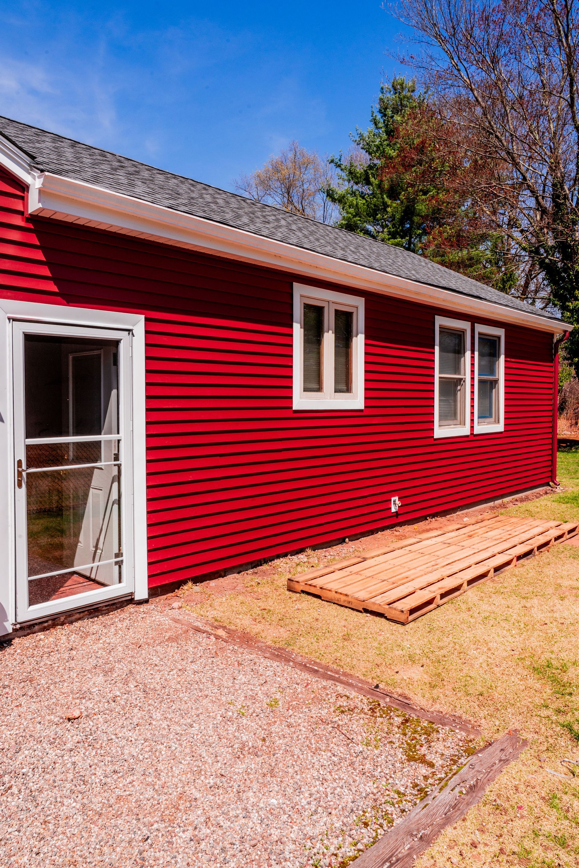 Red-sided house with white-framed windows and door, sitting on a gravel patio under a blue sky.