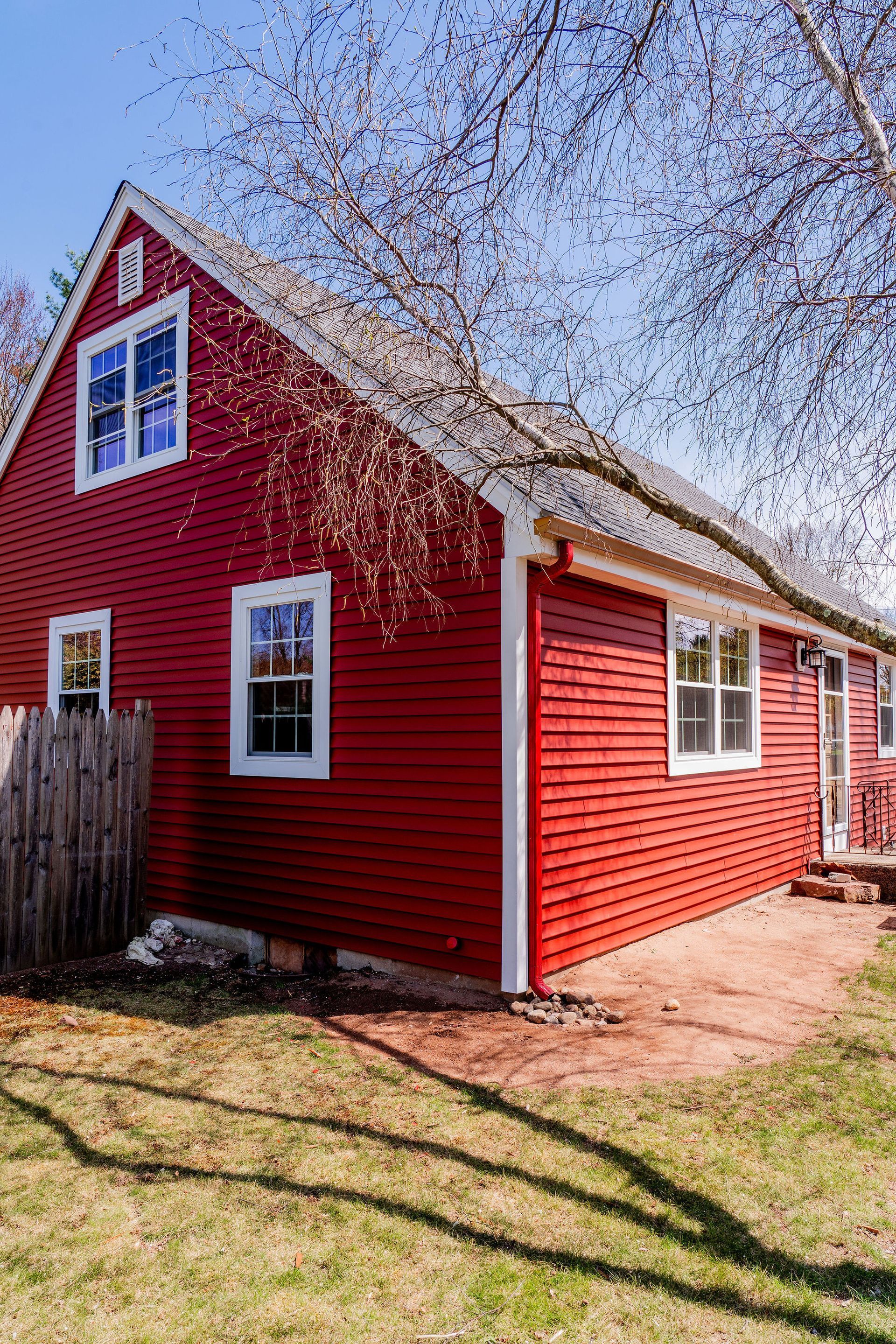 Red house with white trim, a wooden fence, and a tree with budding branches on a sunny day.