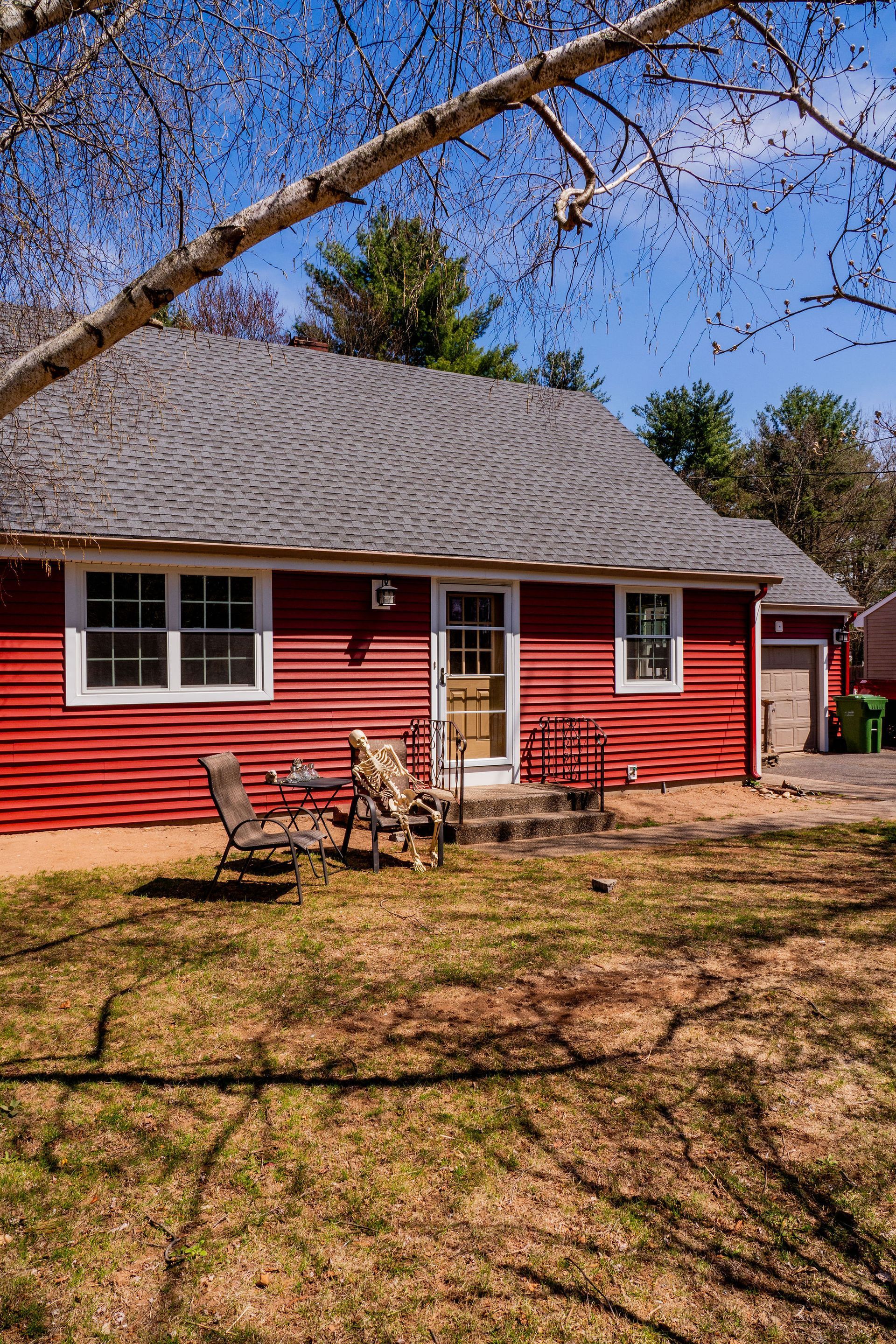 Red house with a gray roof, white-trimmed windows, and two chairs on the lawn under a blue sky.