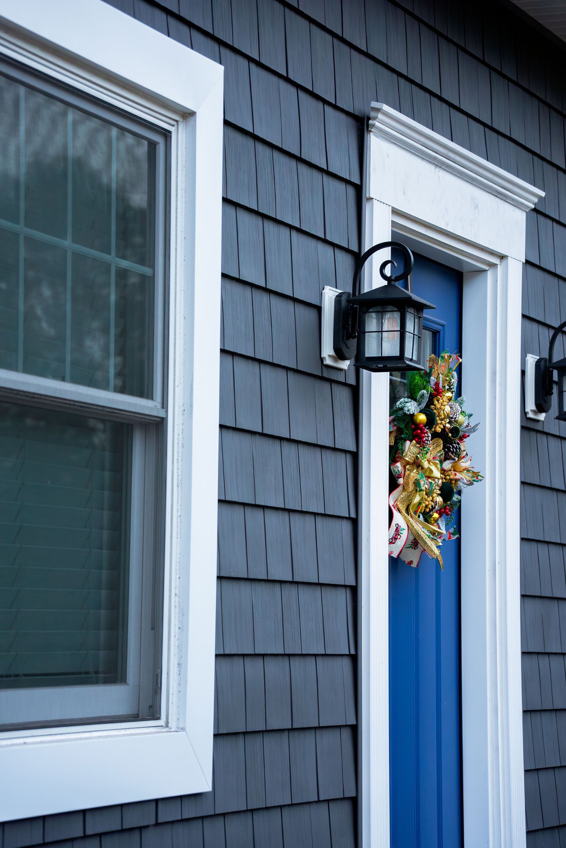 Blue door with a wreath and white trim, gray shingled siding, and a window.