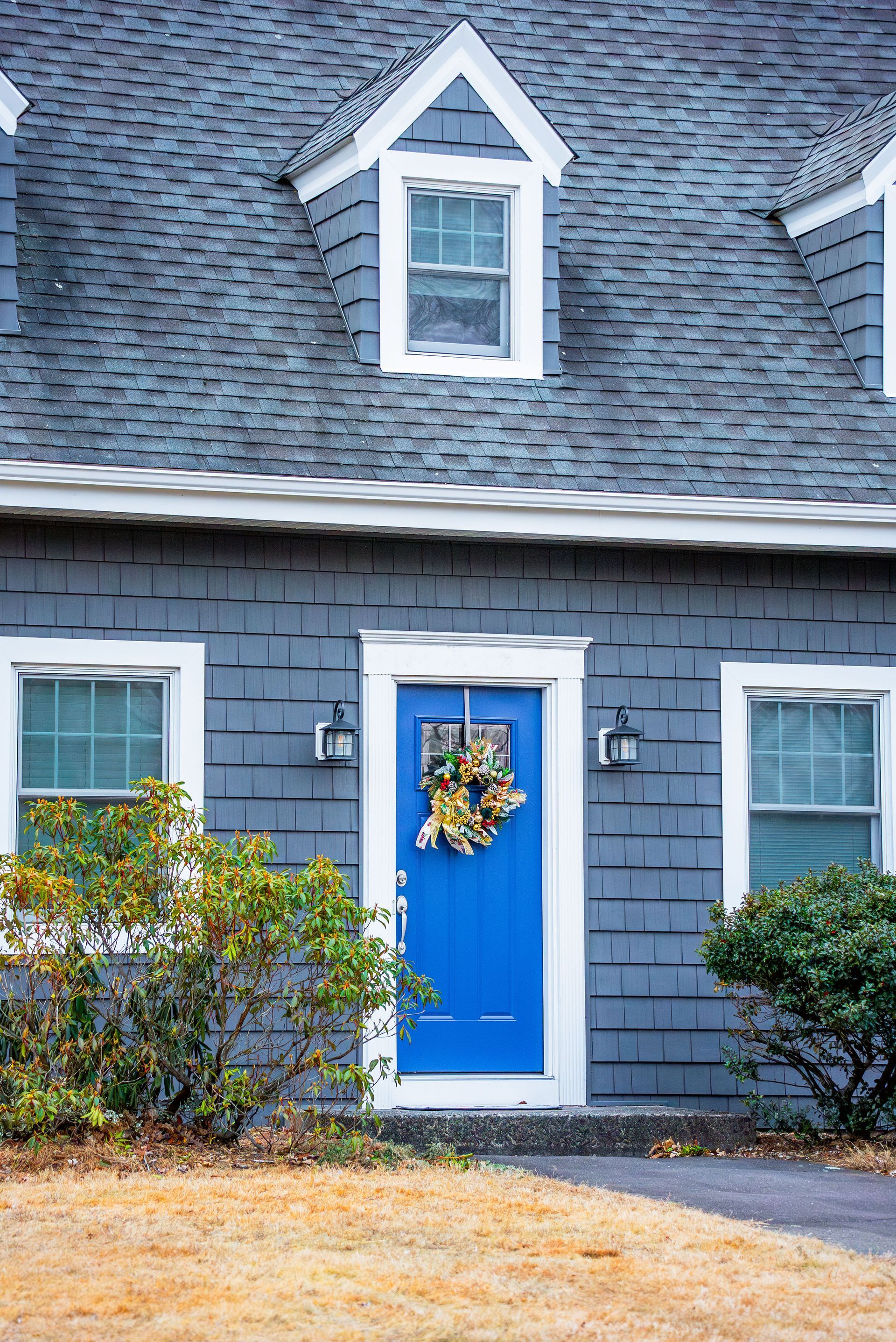 Blue door with gold wreath in a gray shingled house, with white trim and windows.