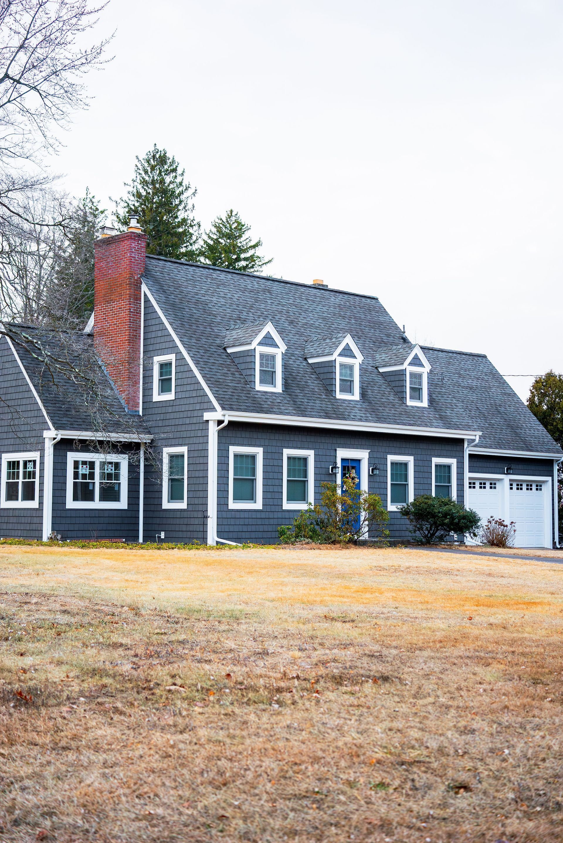 Gray house with white trim, blue door, and a red brick chimney on a cloudy day.