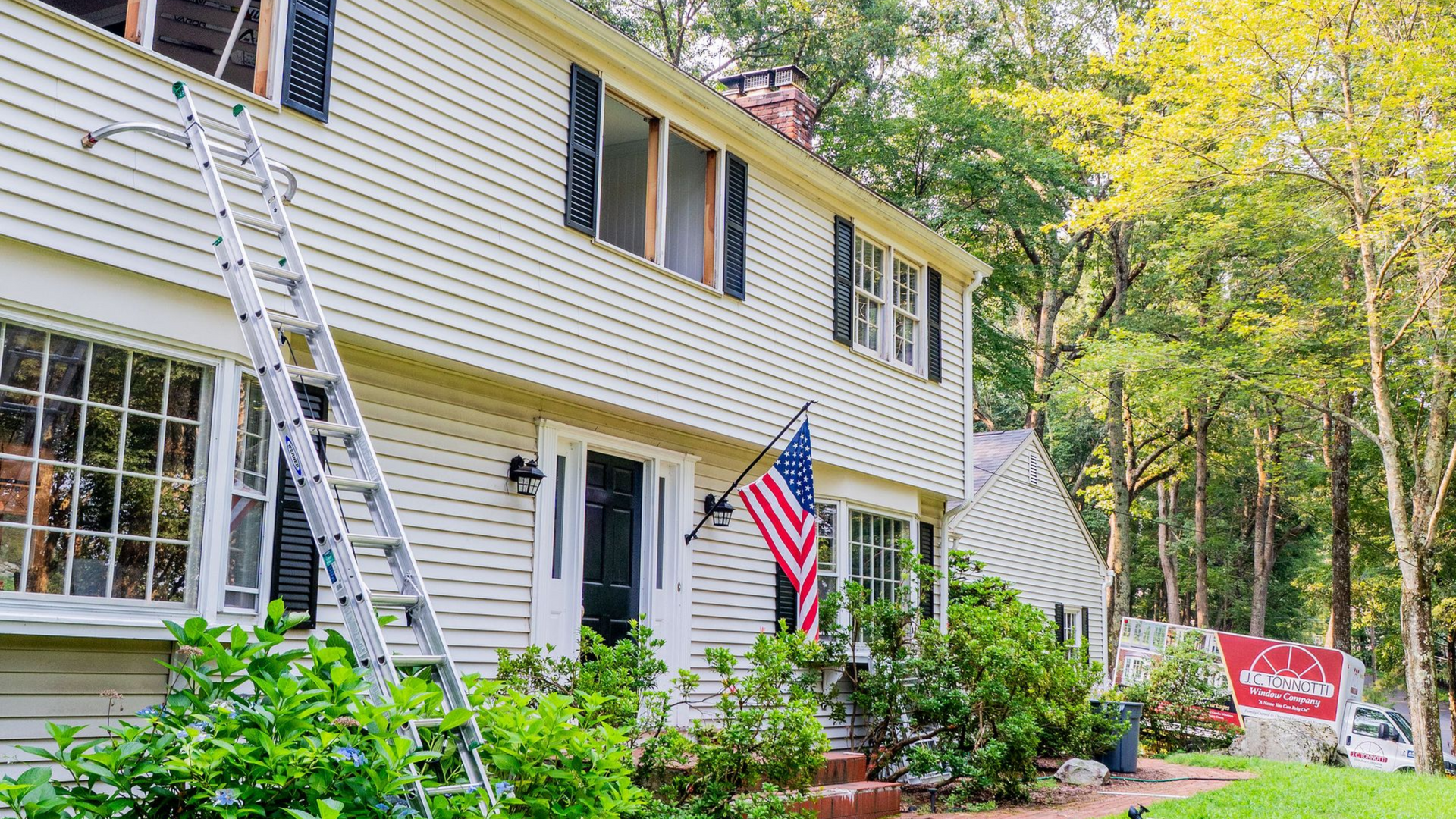 Two-story house with ladder, window replacement in progress. American flag on porch, surrounded by greenery.