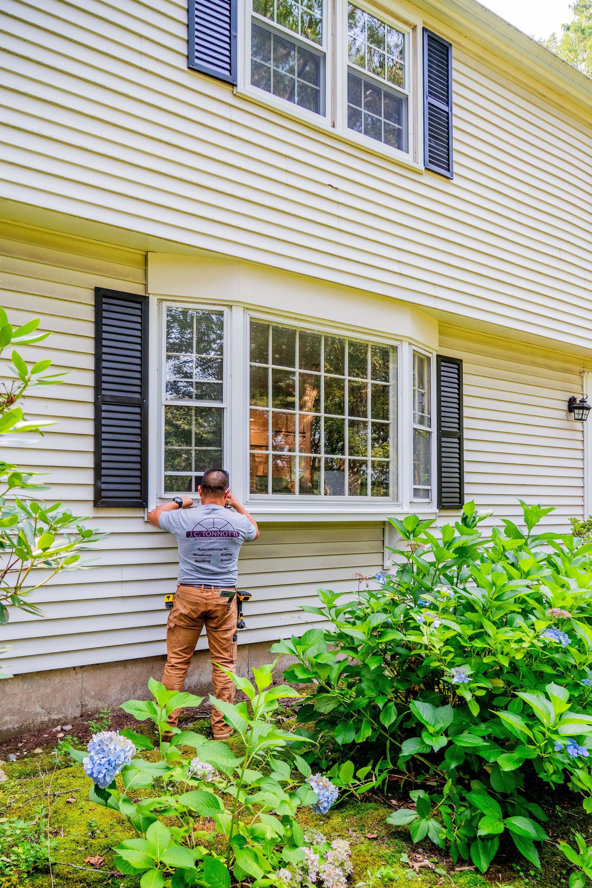 Man installing window, yellow house, black shutters, surrounded by green bushes.