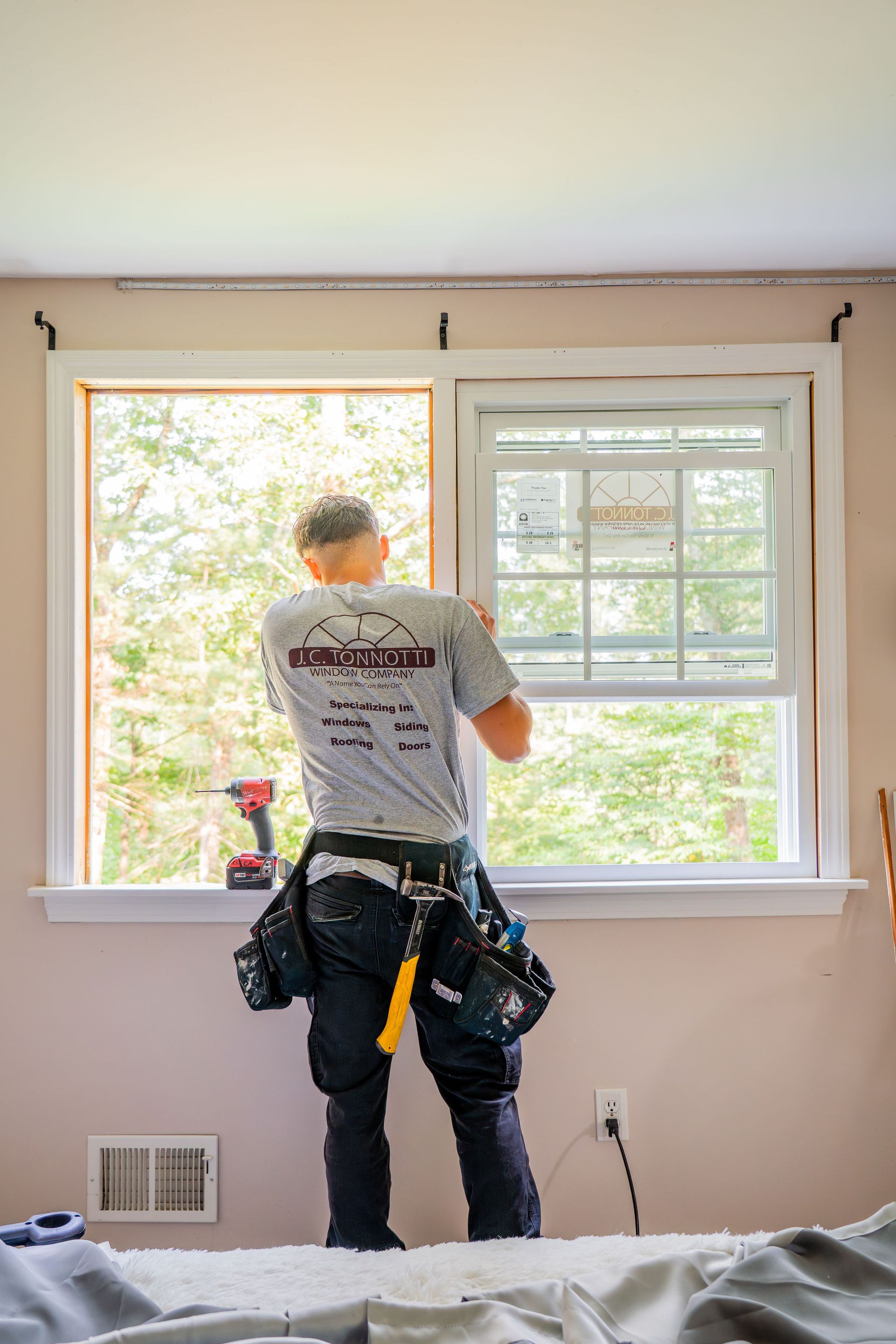 A person installing a window, standing inside a room with tool belt, next to the open window frame.