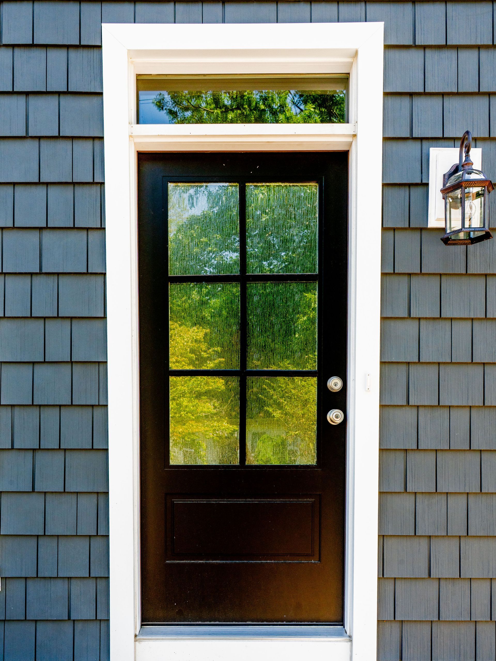 Black door with glass panes, white trim, and a small window above, set in gray shingle siding.