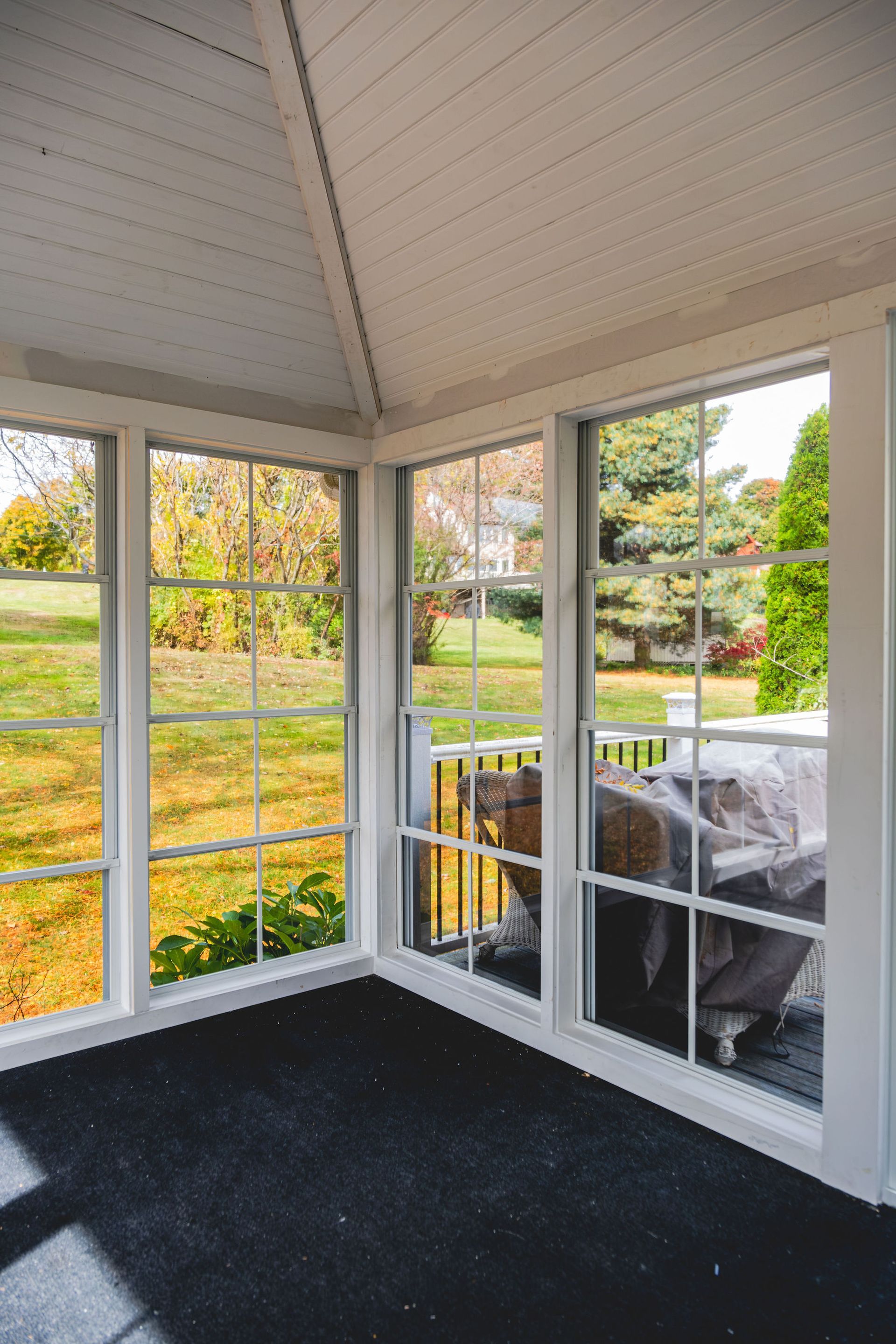 Sunroom with large windows overlooking a backyard. Black carpet, white trim, and autumn foliage.