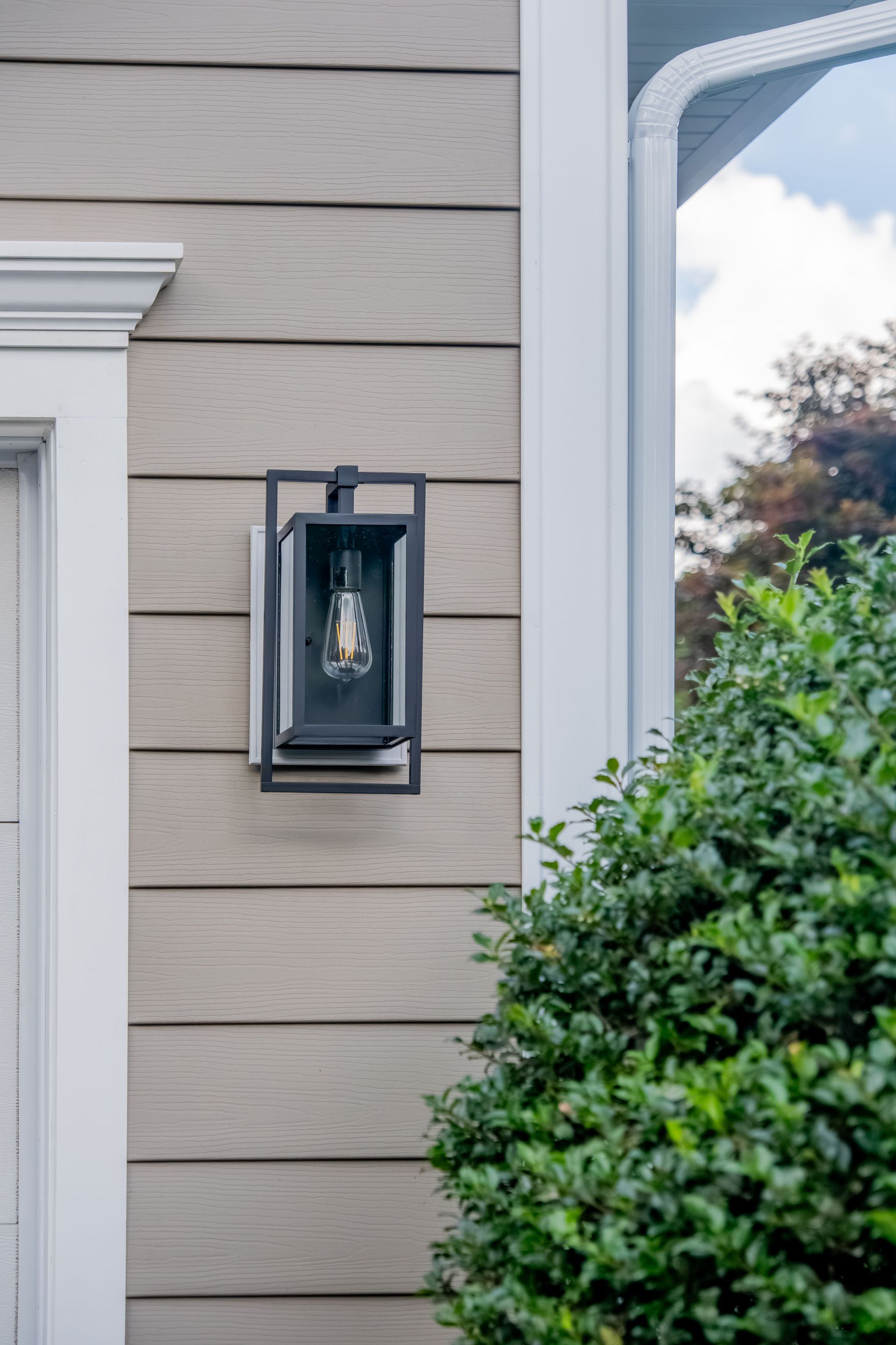 Black outdoor light fixture on tan siding, next to white trim and green bush.