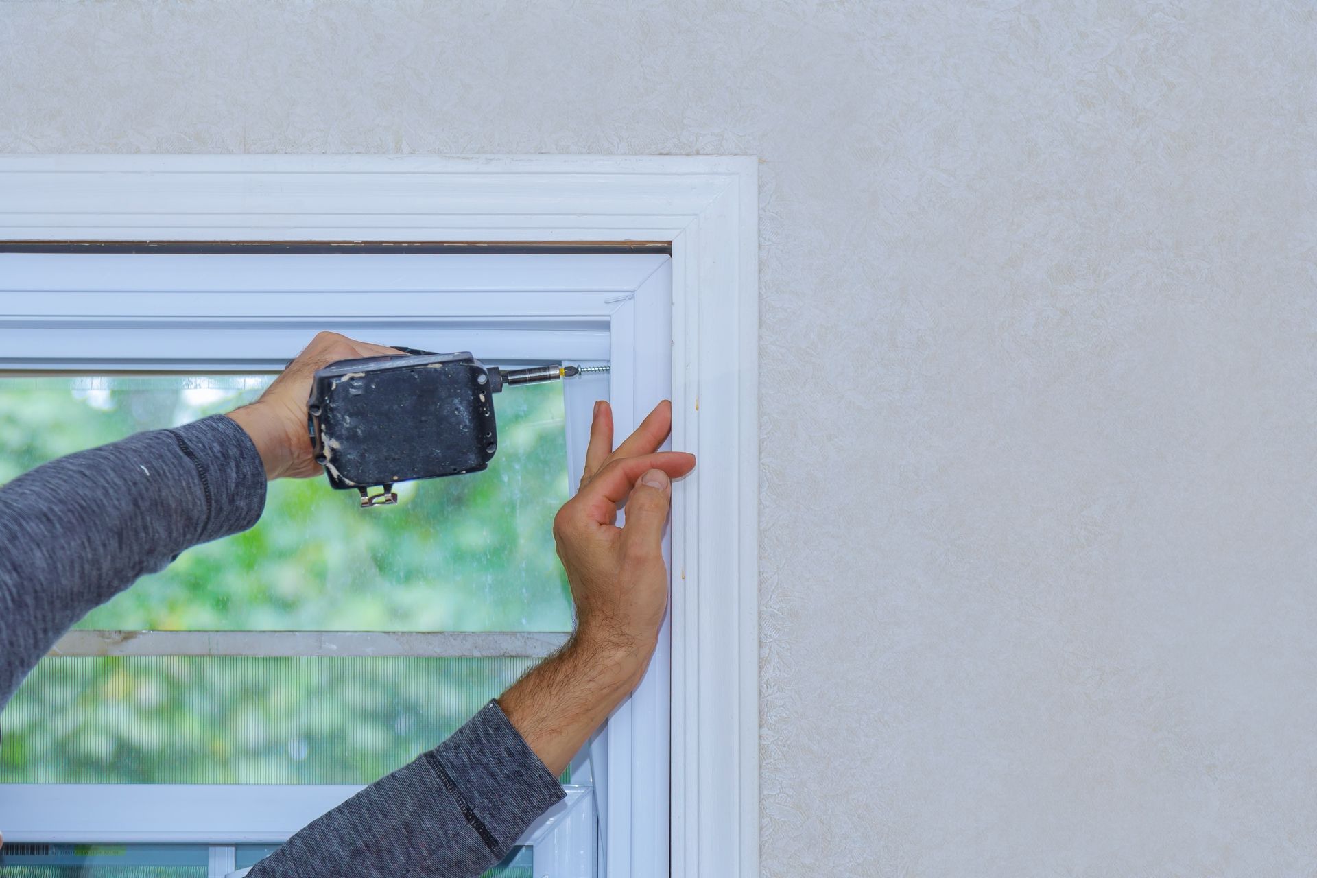 Person using a nail gun to install white trim around a window on a stucco wall.