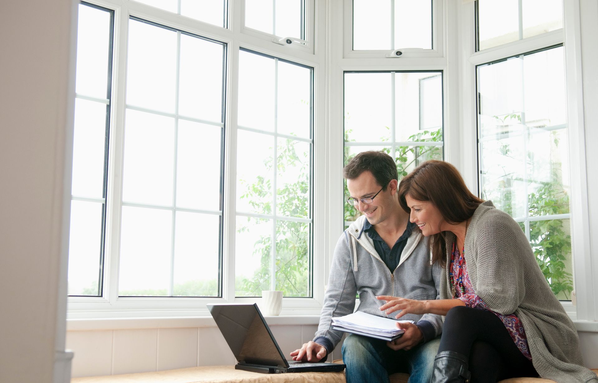 A couple sits in a window seat looking at a laptop and documents, smiling while collaborating in a sunlit room.