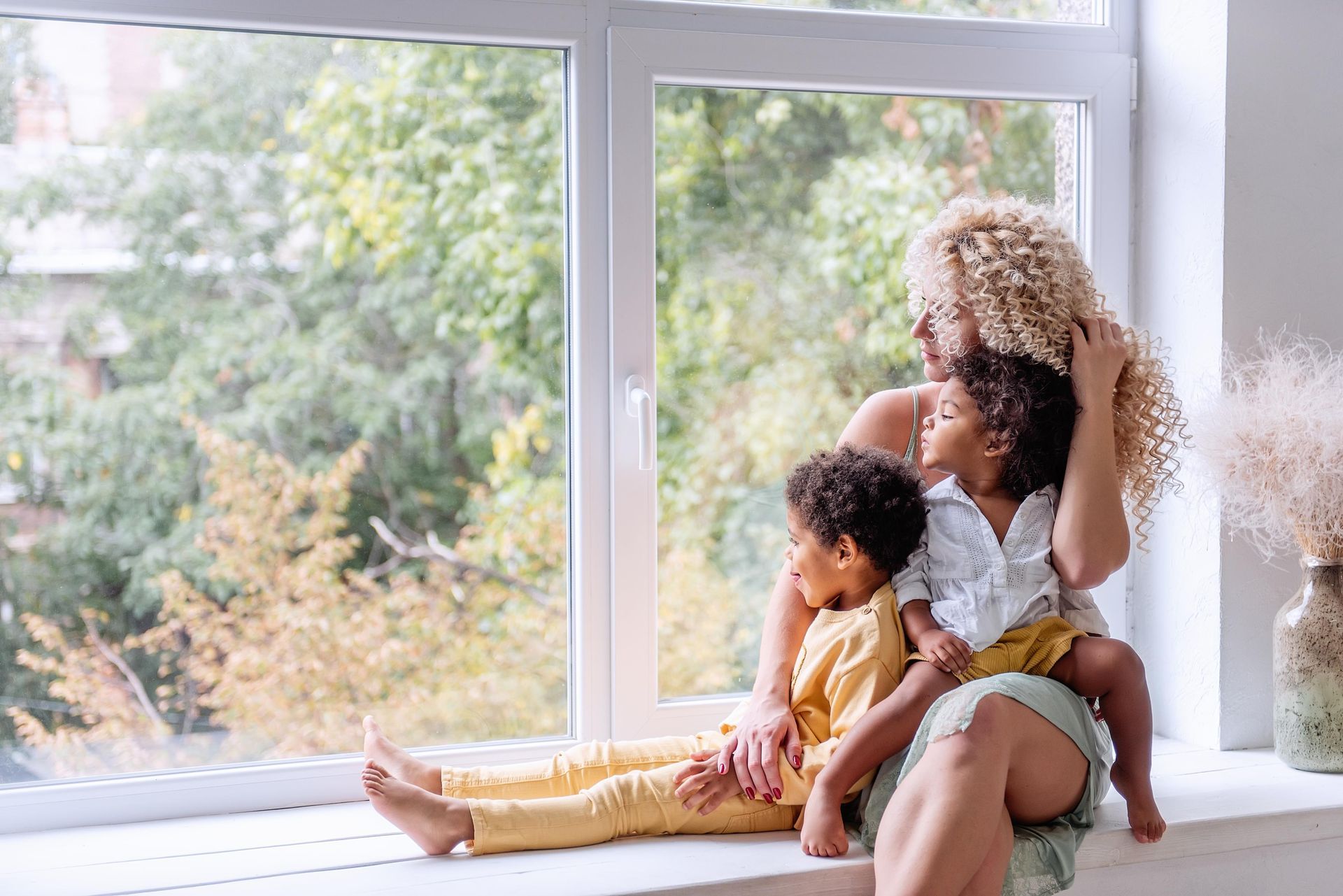 Woman and two children sit on a windowsill, looking out at trees.