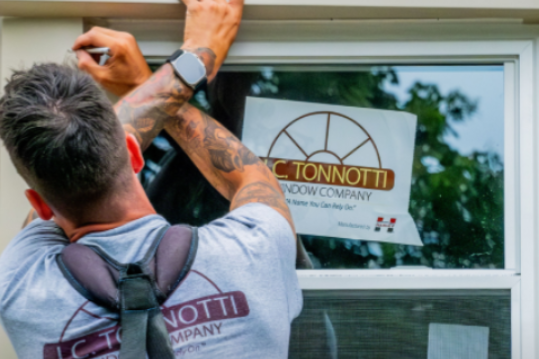 Person installing a window, wearing a company shirt with a logo, in a building.