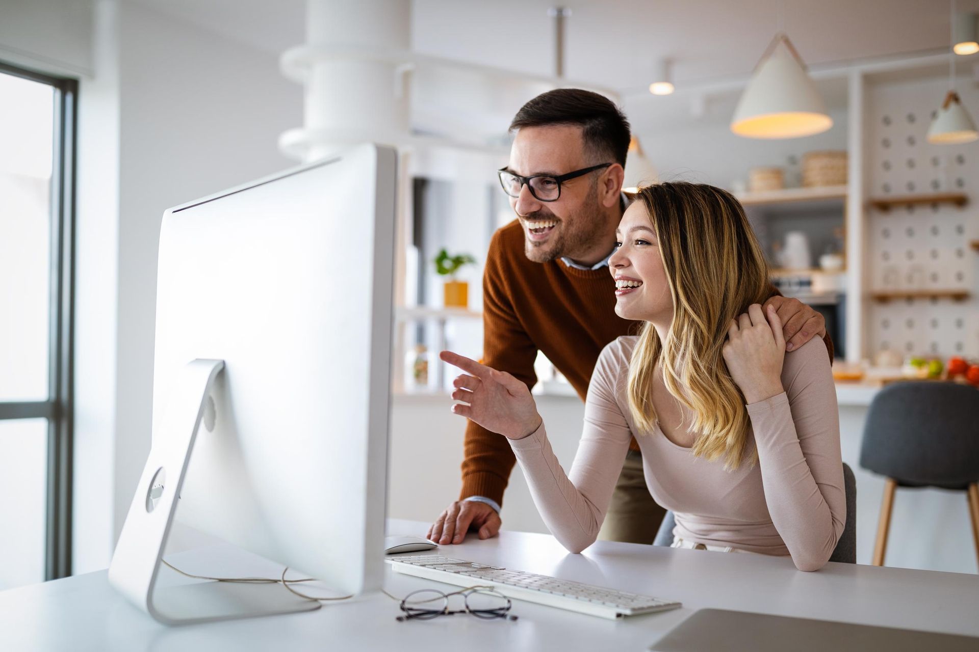Couple looking at computer screen, woman pointing, smiling, man wearing glasses, kitchen background.
