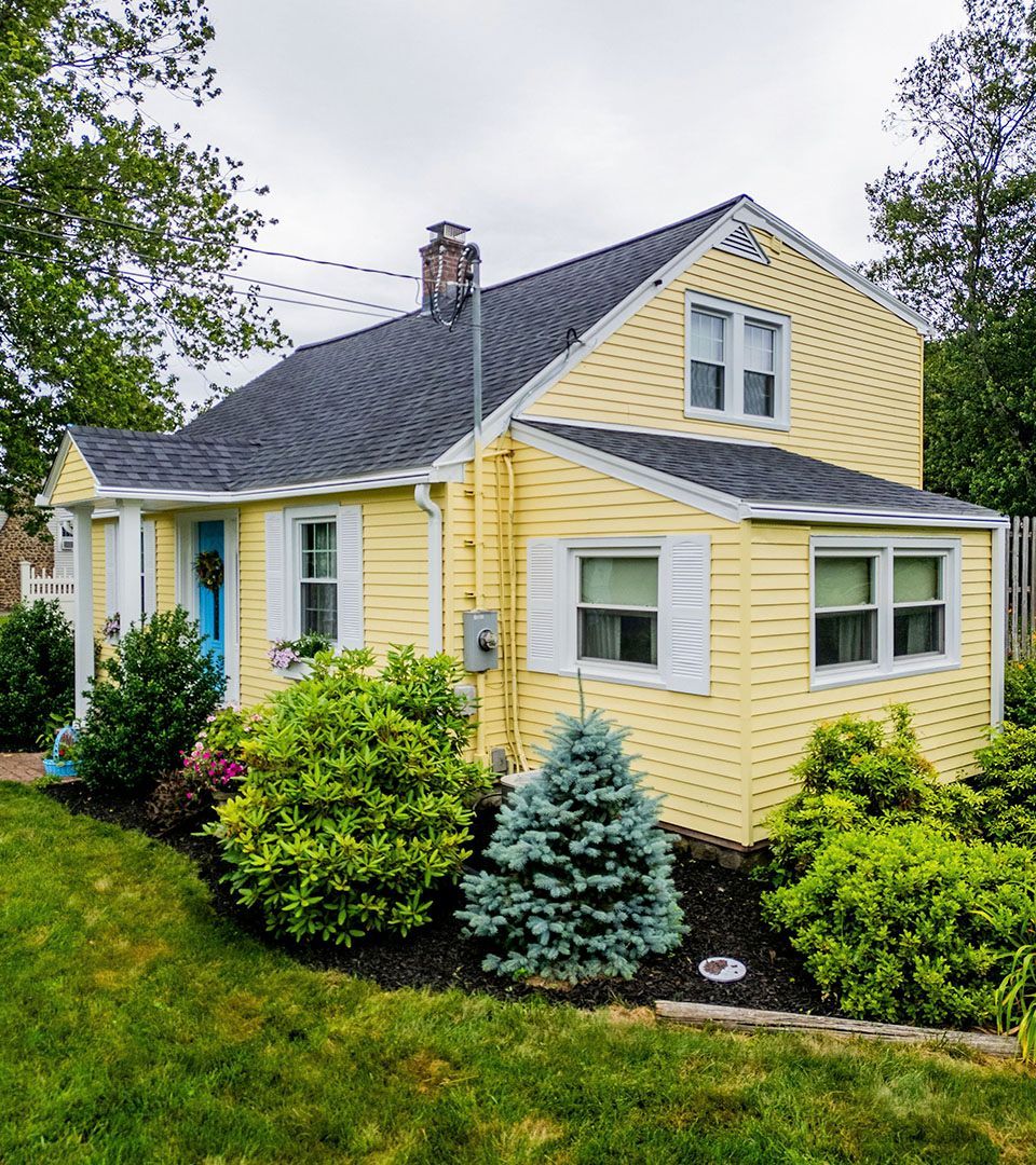 A yellow house with a black roof and white shutters is surrounded by trees and bushes.