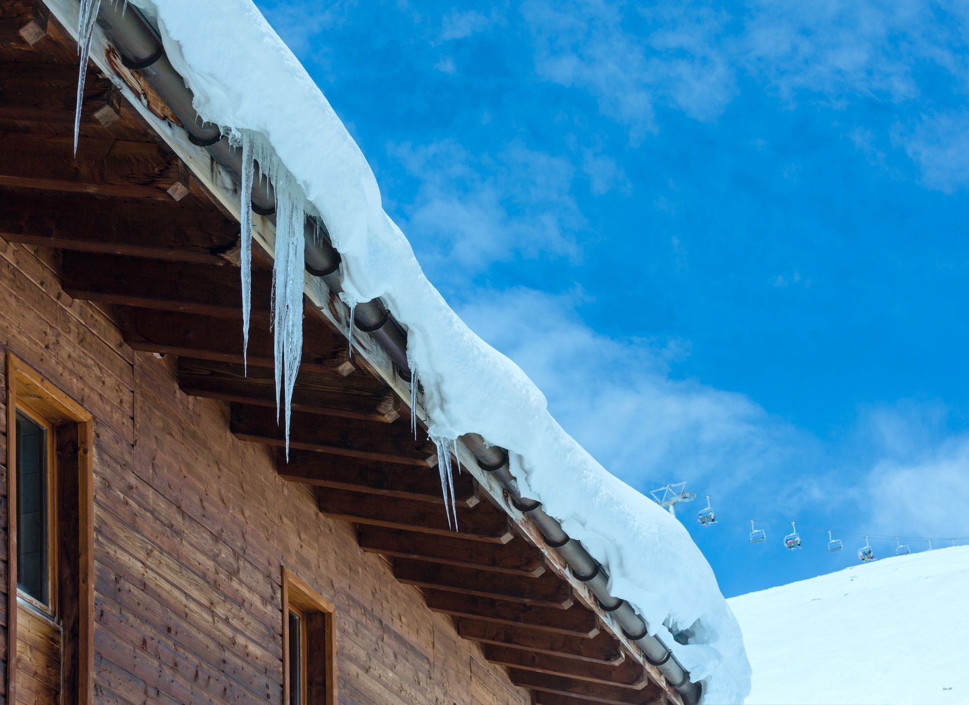 Snow-covered roof with icicles hangs over a wooden building against a bright blue sky.