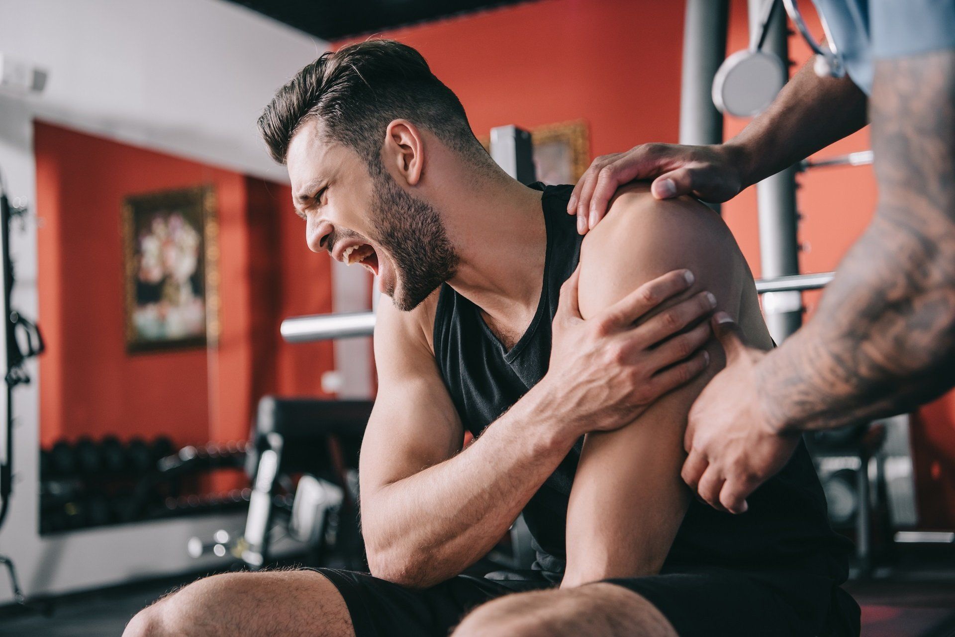 A man is sitting on the floor in a gym holding his shoulder in pain.