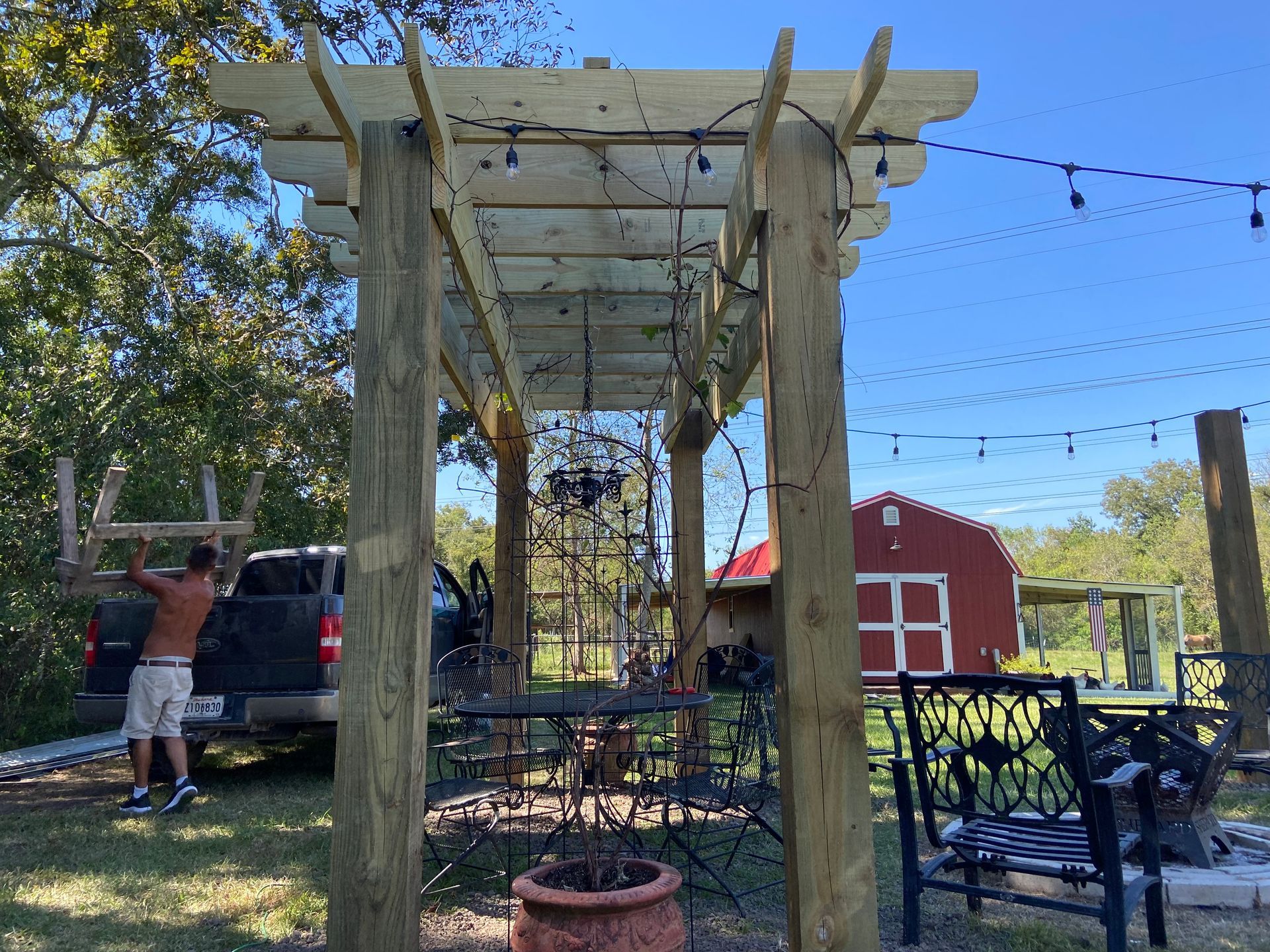 A man is standing under a wooden pergola in front of a red barn.