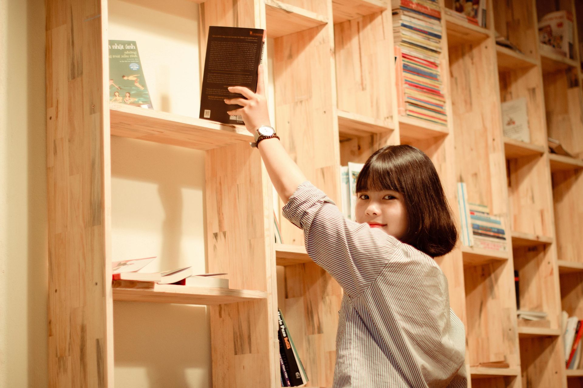A woman is reaching for a book on a wooden shelf.