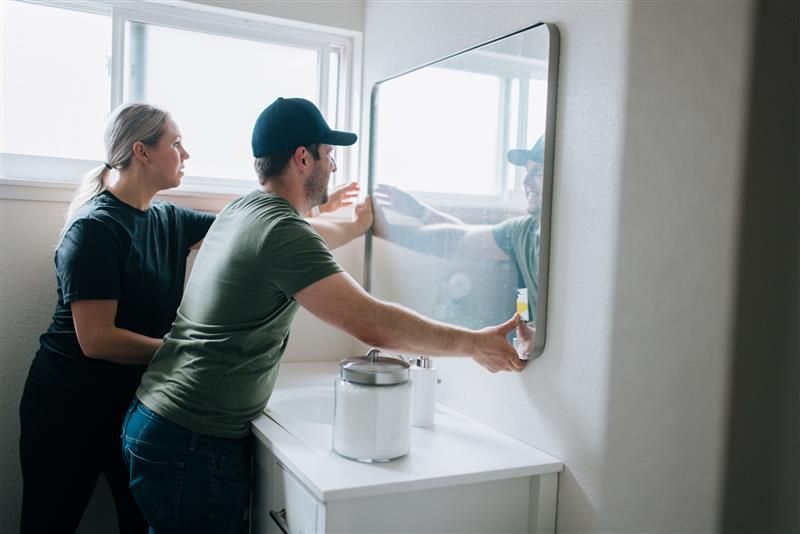 A couple places a new mirror on a bathroom wall.