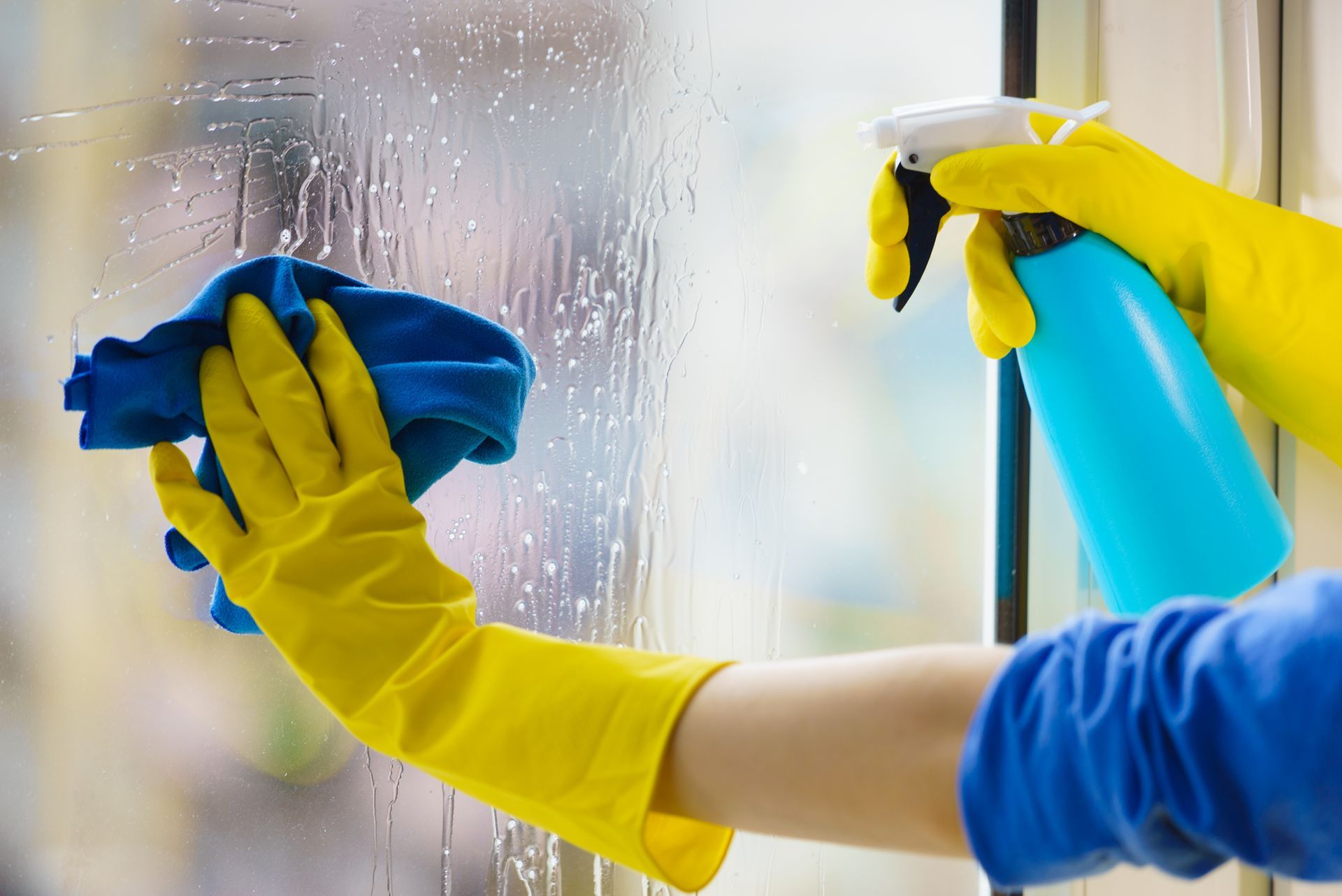 Gloved hands cleaning a foggy window with a blue cloth and spray bottle